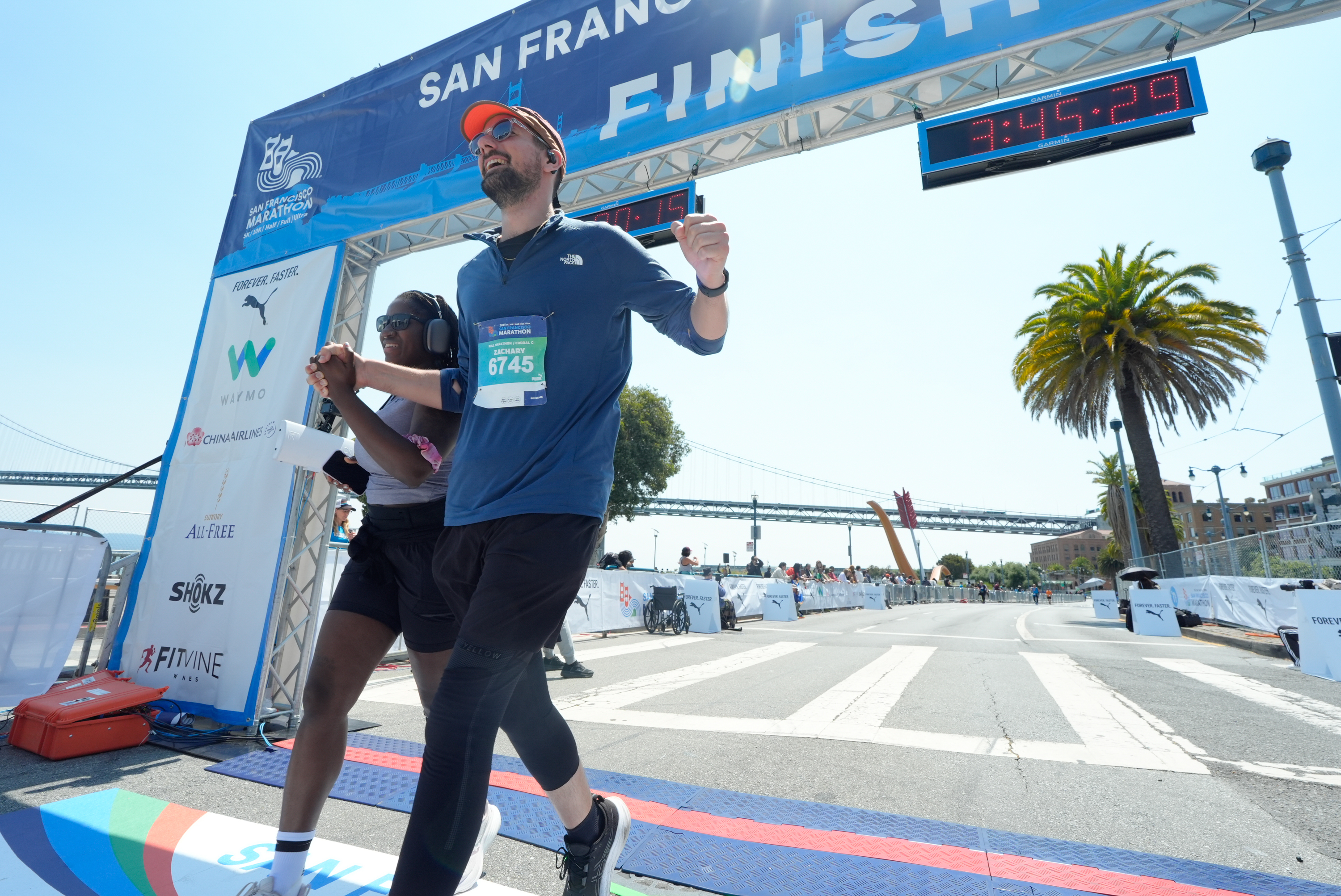 Two runners holding hands cross the finish line of the San Francisco Marathon, smiling. The race clock reads 3:45:29, and a bridge and spectators are visible in the background.