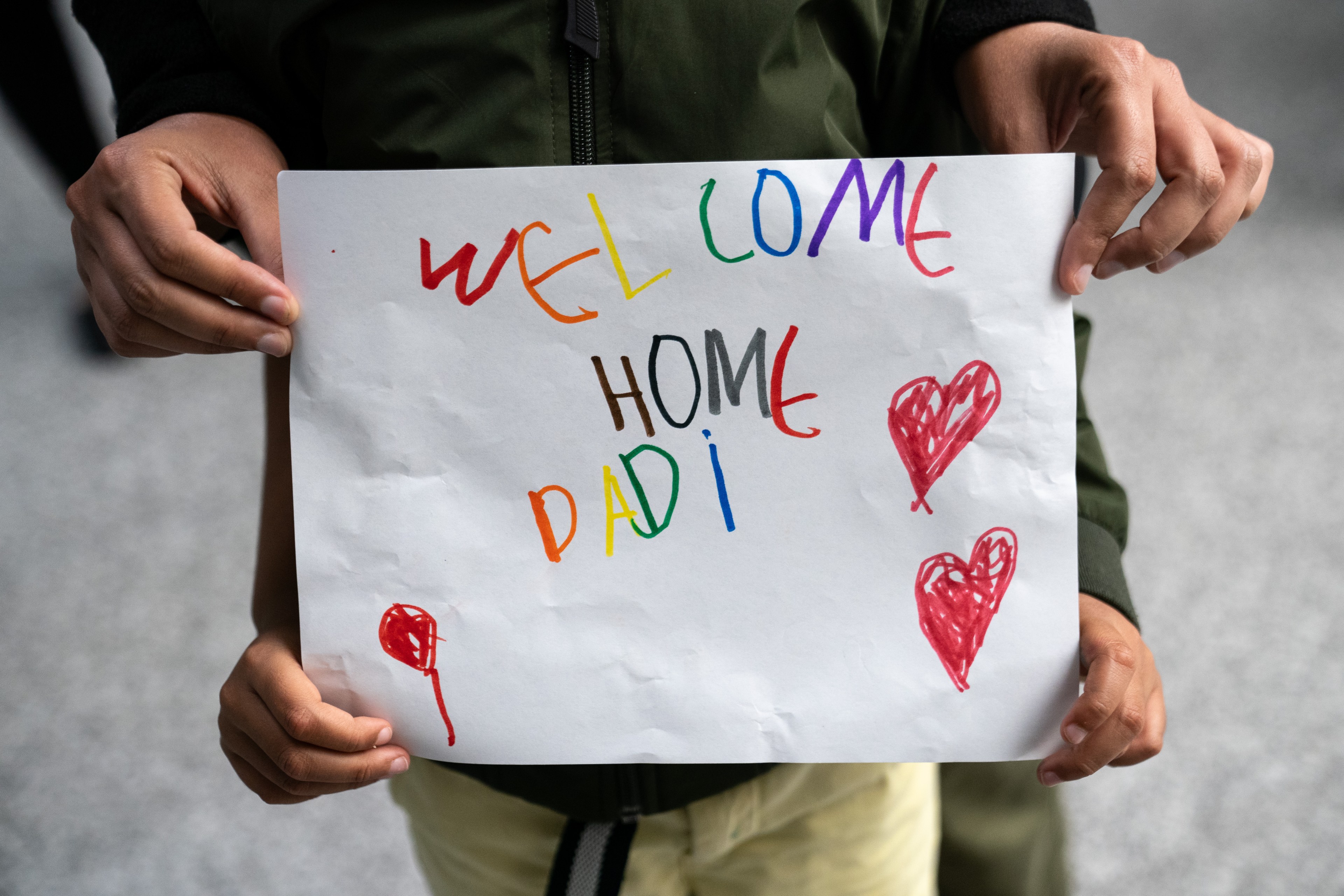 A child holds a hand-drawn sign saying &quot;Welcome Home Dadi&quot; in colorful letters, decorated with three red hearts. An adult's hands are visible supporting the child’s hands.