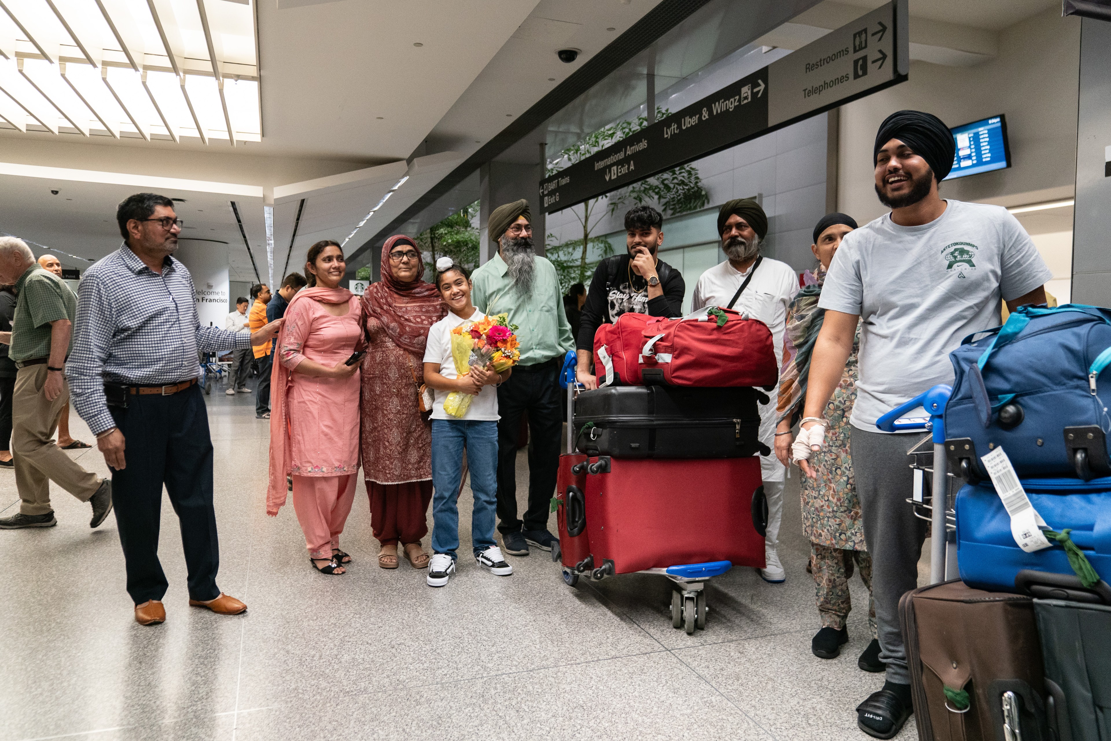 A group of people, including women, men, and a child holding flowers, stand in an airport arrivals area with luggage, under signage for restrooms and rideshare options.