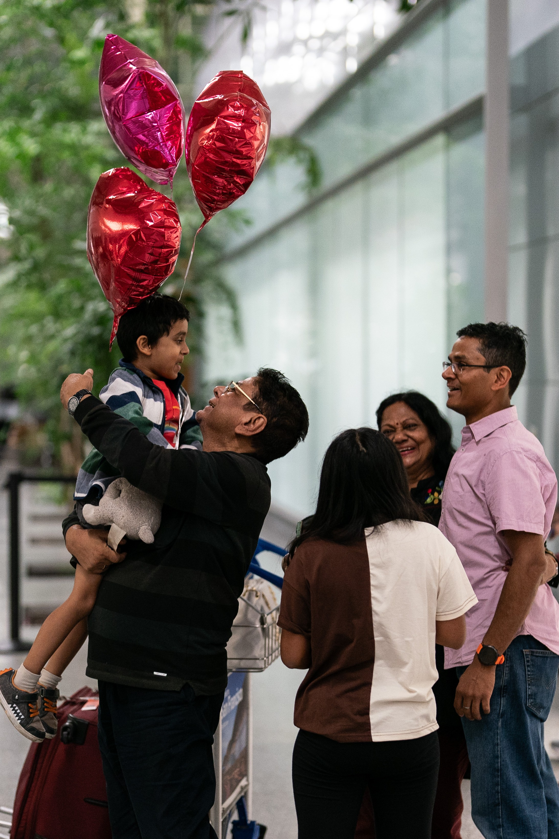 A group of people is at an airport greeting a mother and child, who is being lifted and holding red heart-shaped balloons. Luggage and travelers surround them.