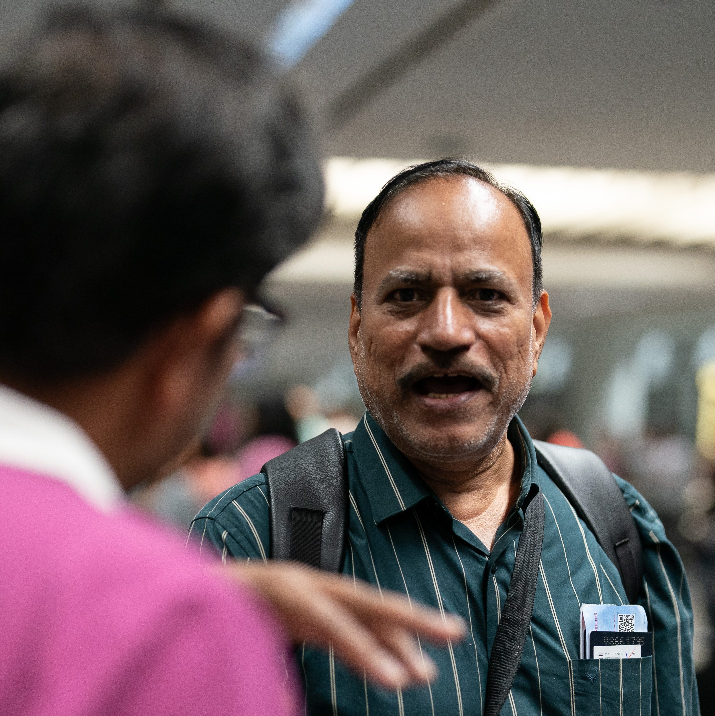 Two people are engaged in a conversation indoors. One man is in focus, wearing a green striped shirt, carrying a backpack, and holding travel documents. The other man is blurred and seen from behind.