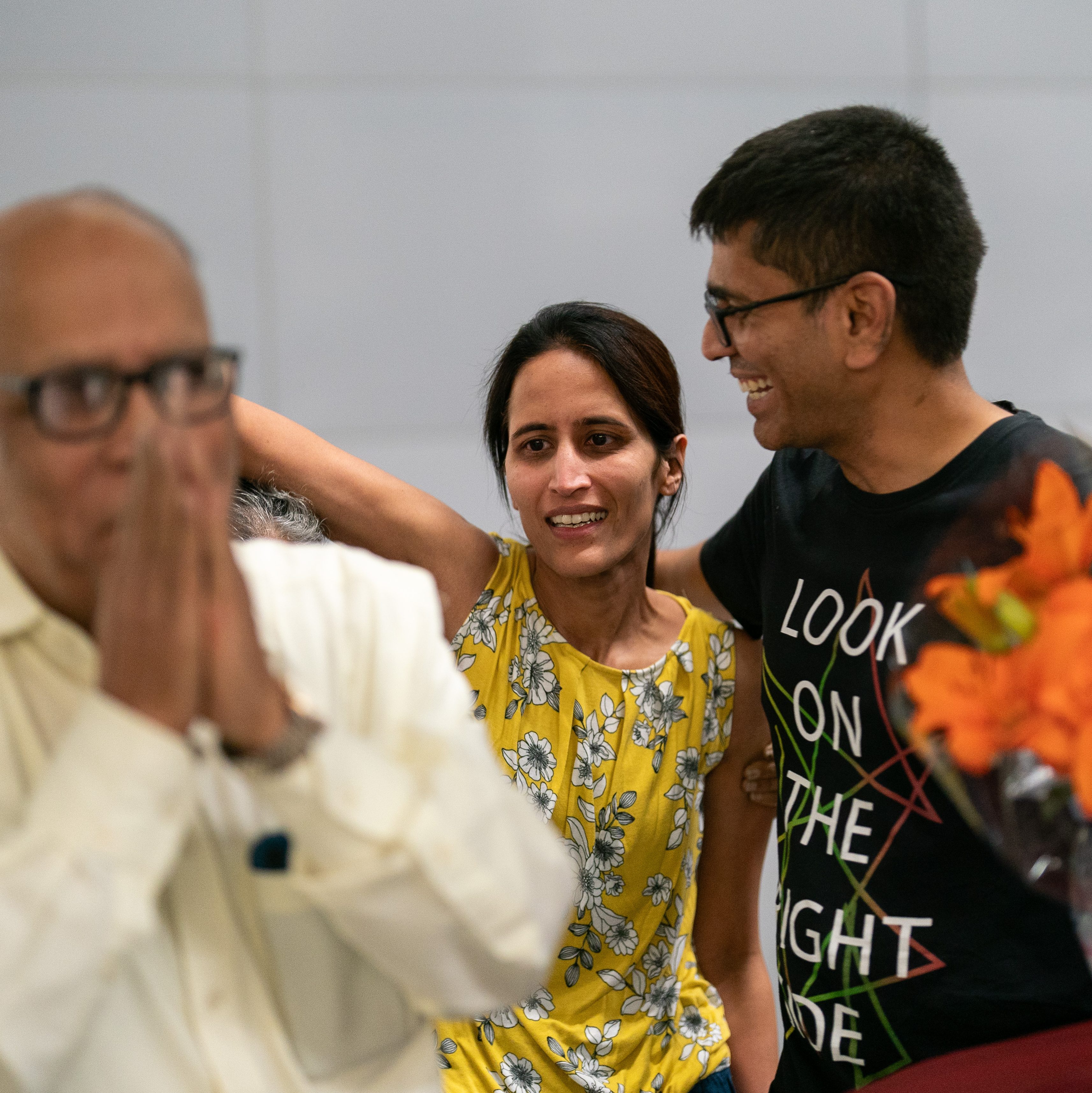 An elderly man in a white shirt stands with hands clasped in front of his face, while a woman in a yellow floral top and a man holding flowers smile and embrace.