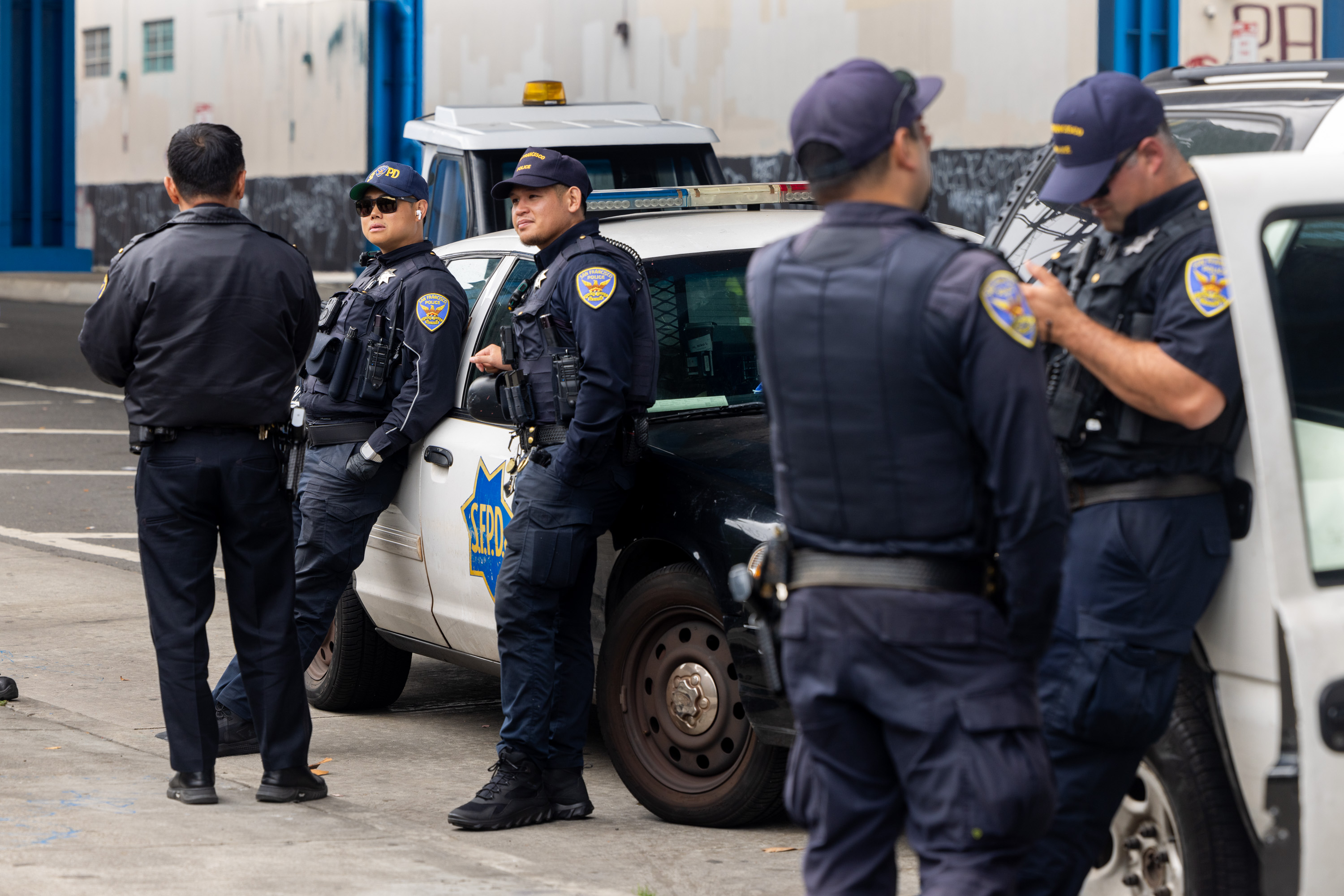 A group of police officers stand and talk near a patrol car outside a building. Some officers lean on the car, while one reads something from a paper.