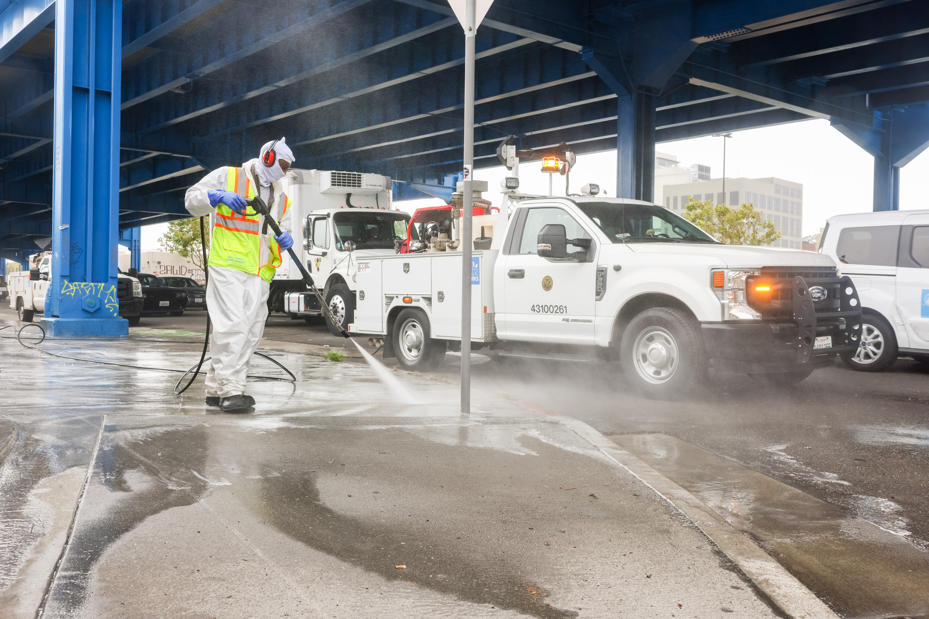 A worker in protective gear and a high-visibility vest is power-washing a wet sidewalk under a blue bridge, with utility trucks parked nearby.