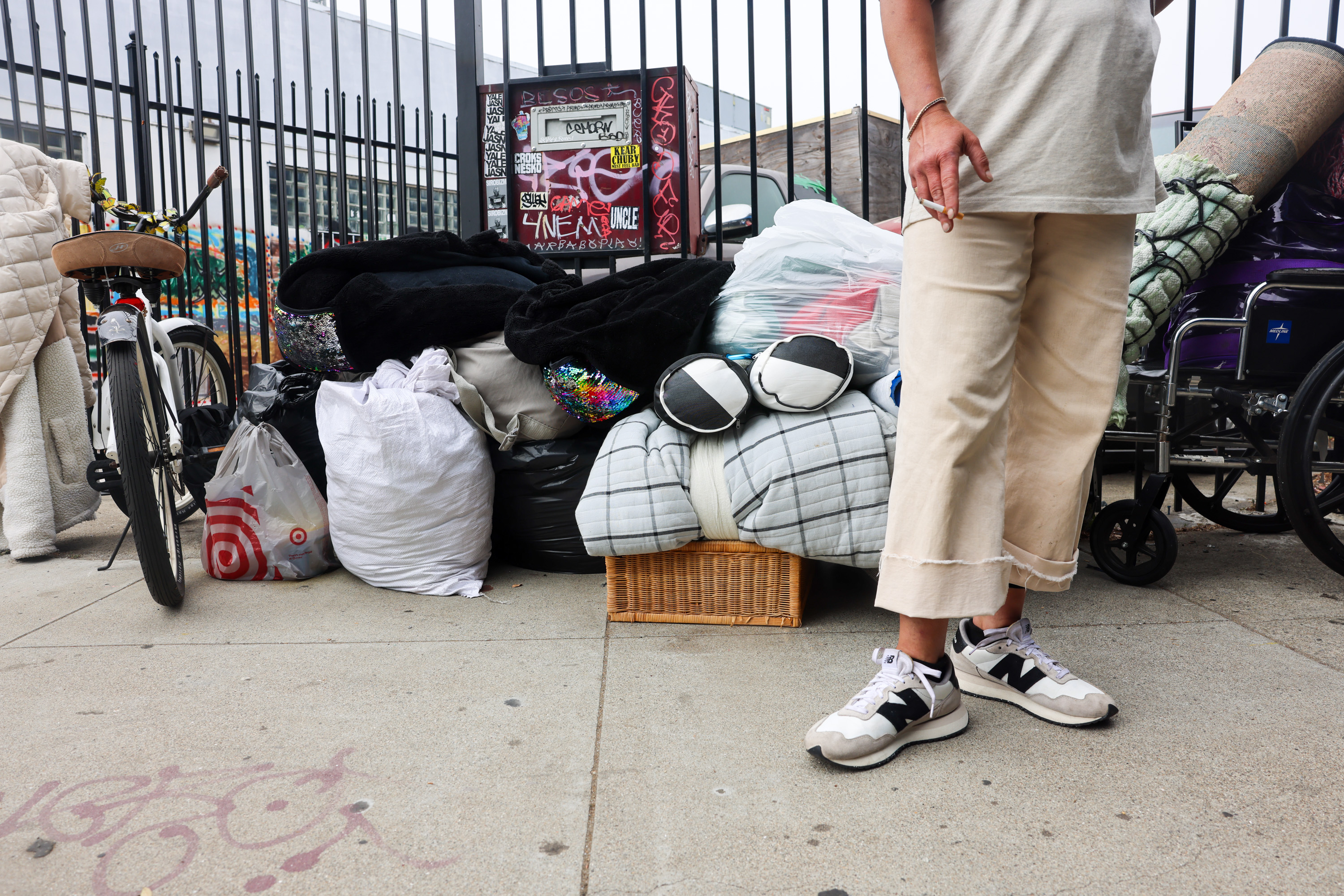 The image shows a person standing next to a pile of bags, blankets, and a bicycle, with grid fencing and graffiti in the background. The person is holding a cigarette.