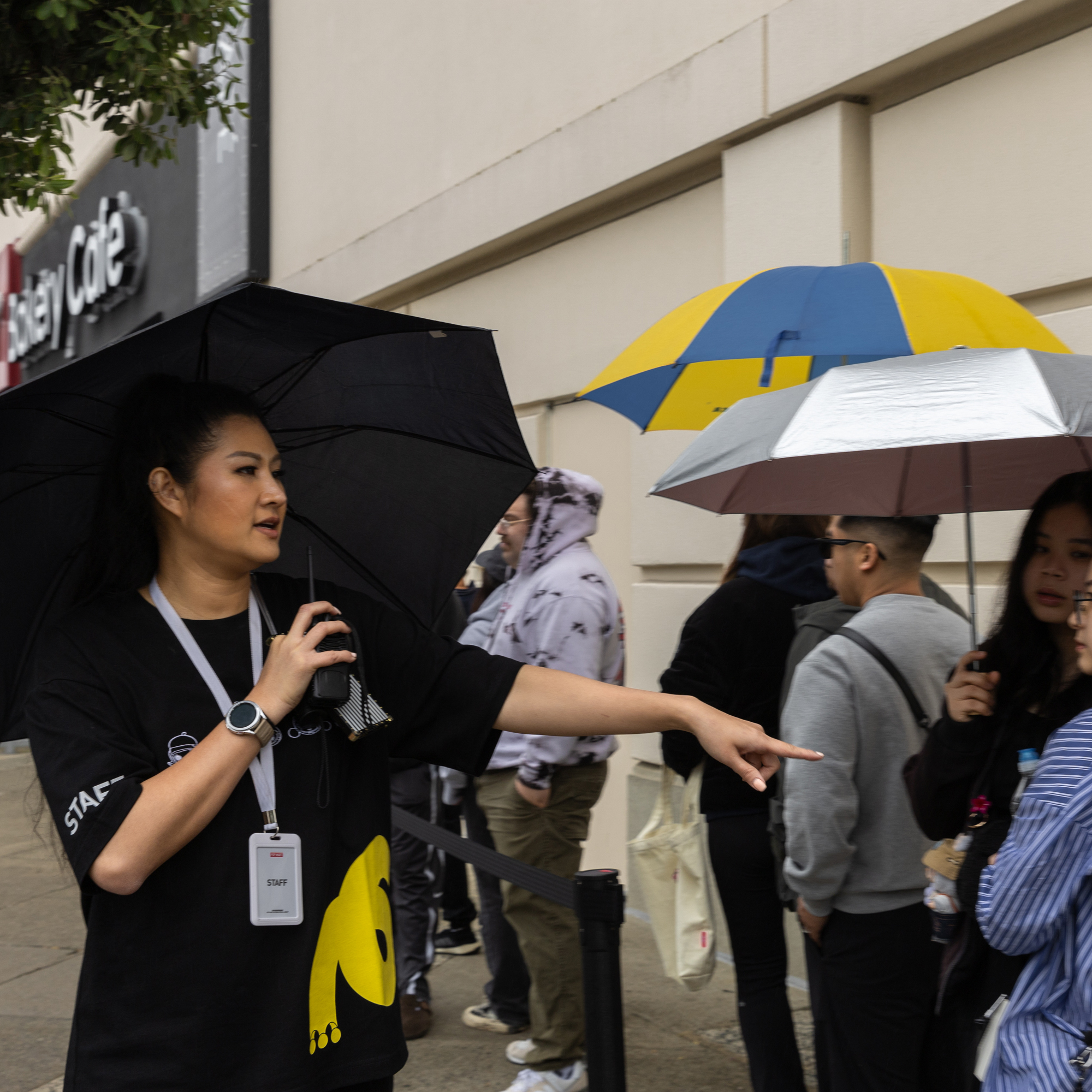 A staff member holding a black umbrella directs a line of people outside; individuals hold umbrellas and wear hoodies, standing near a beige building with a sign.