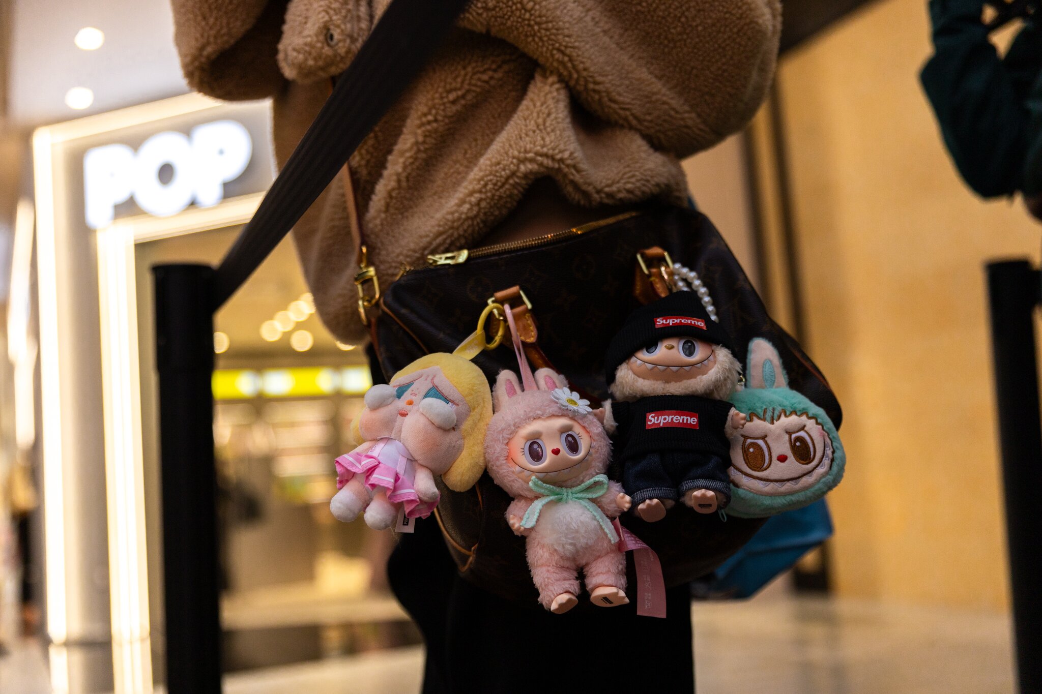 A person with a beige sweater has a Louis Vuitton bag decorated with four cute plush keychains, each with unique outfits and expressions, against a shopping mall backdrop.