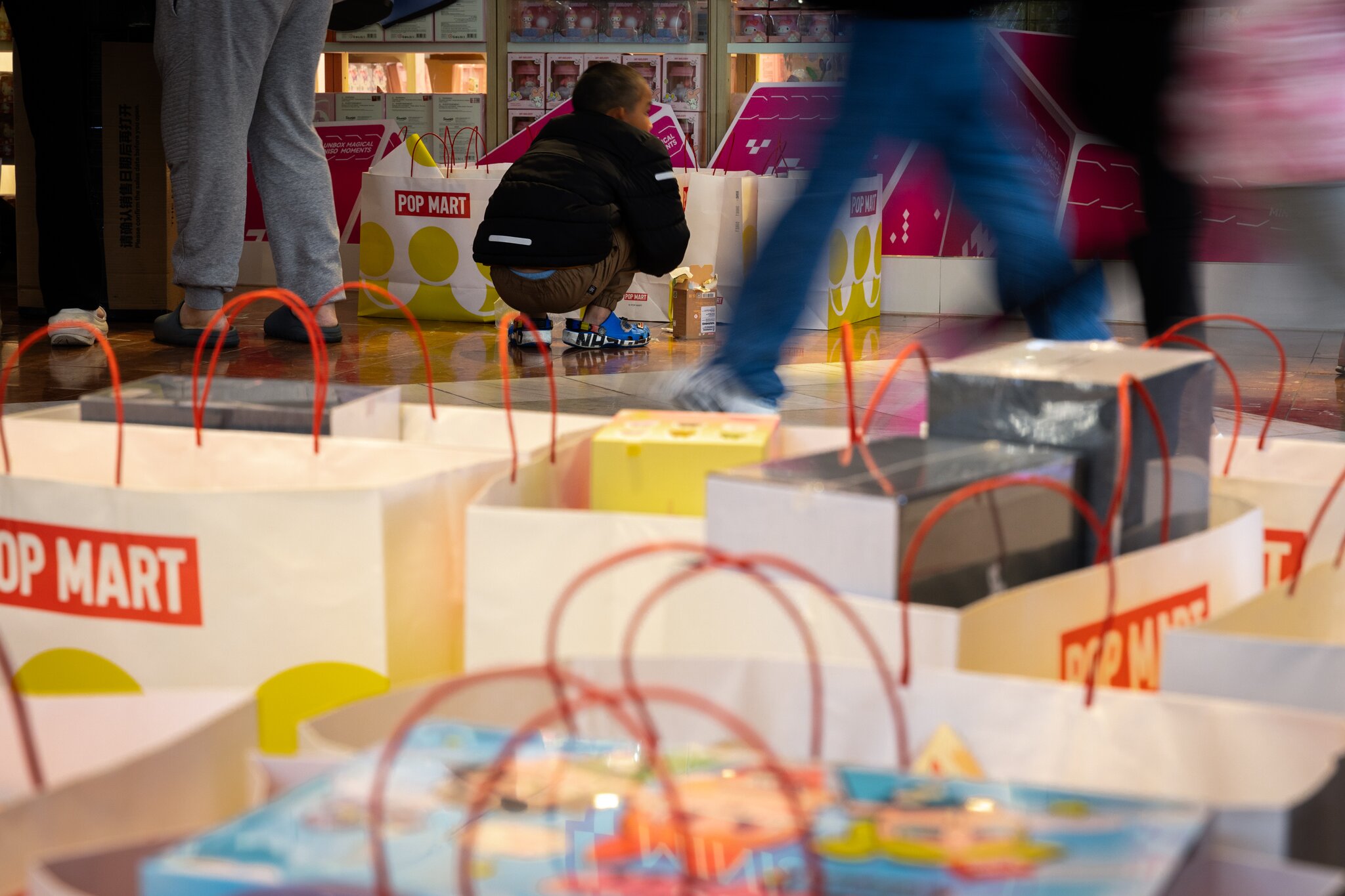 A child squats next to large shopping bags in a store named &quot;Pop Mart,&quot; with people standing and walking around in the background.