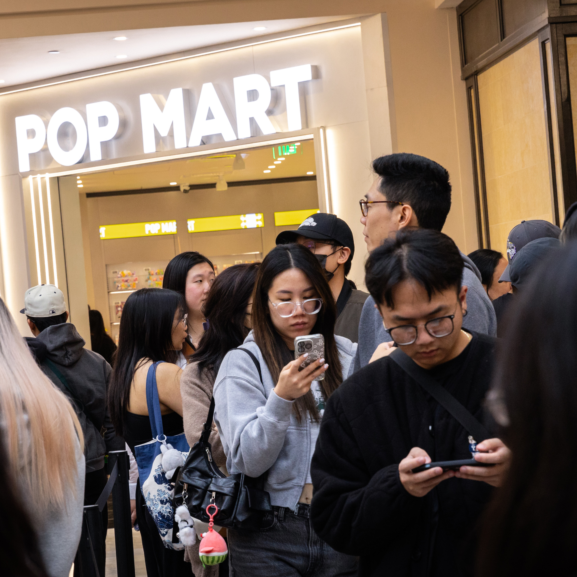 People are gathered in front of a store named &quot;POP MART&quot;, with many looking at their phones while standing in line.