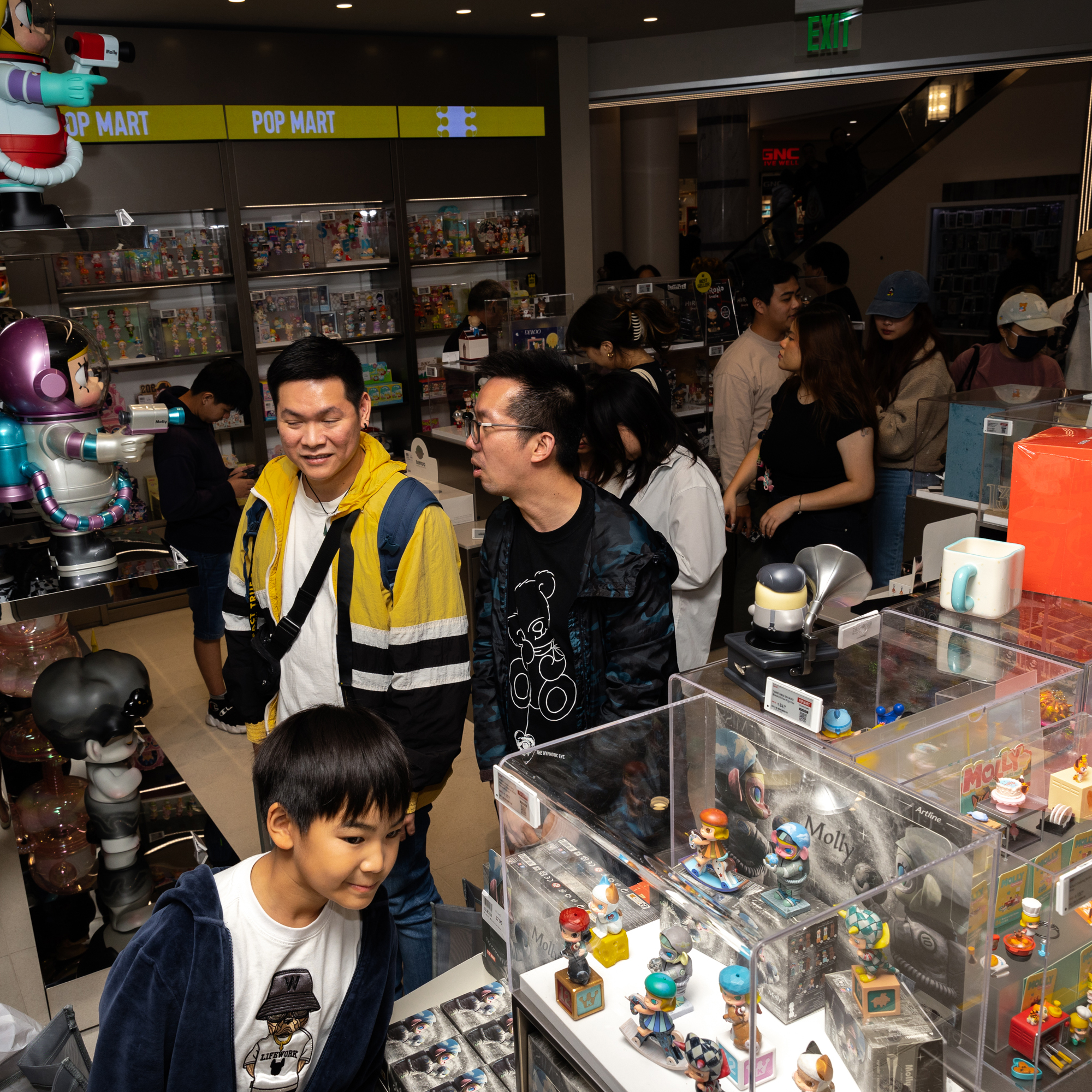 The image shows people browsing a Pop Mart store filled with colorful toy figures. Shelves are lined with various collectibles in bright packaging.