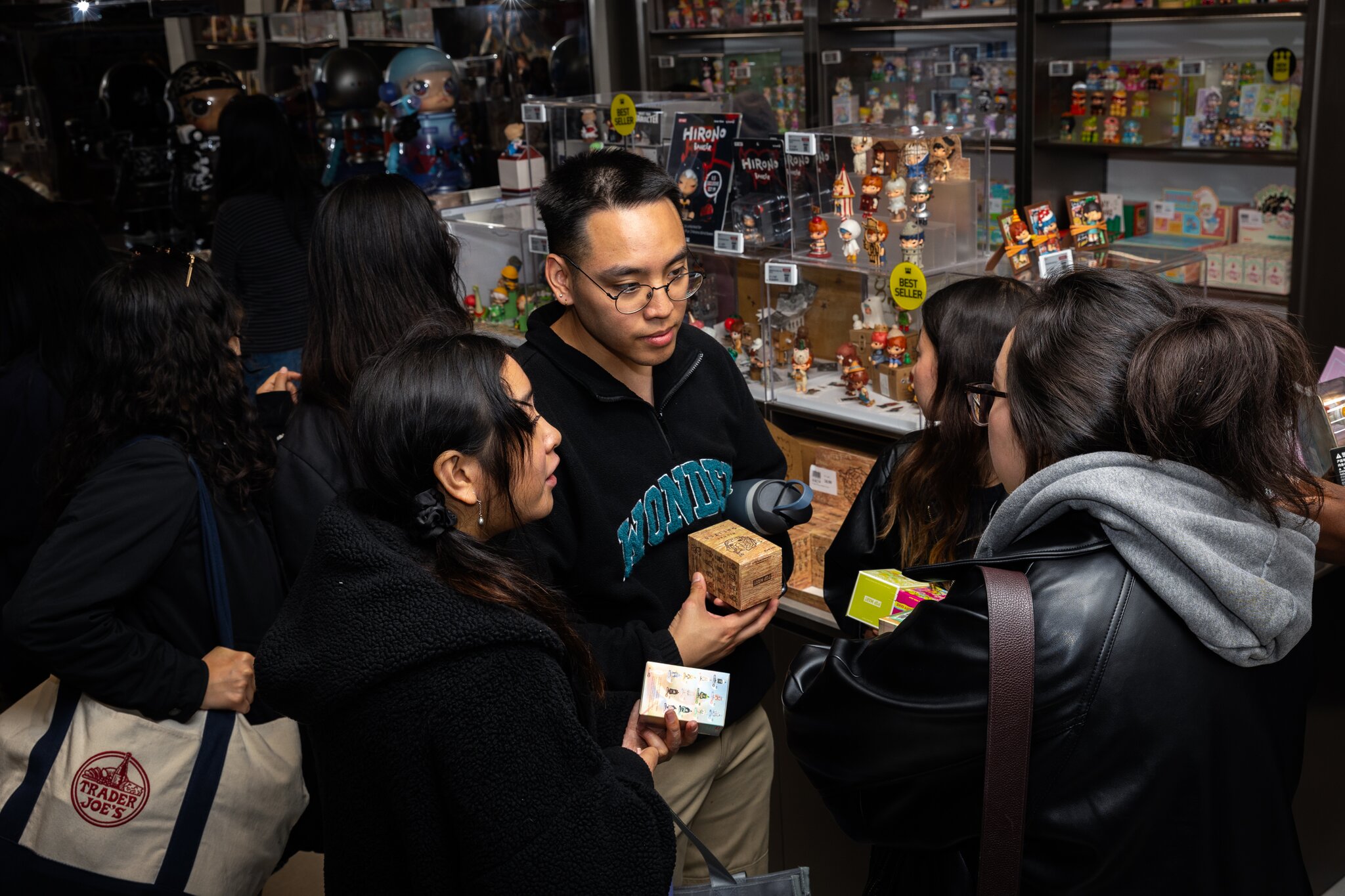 A group of people is gathered in a toy store, holding items and conversing. Shelves filled with various figurines and collectibles are displayed in the background.