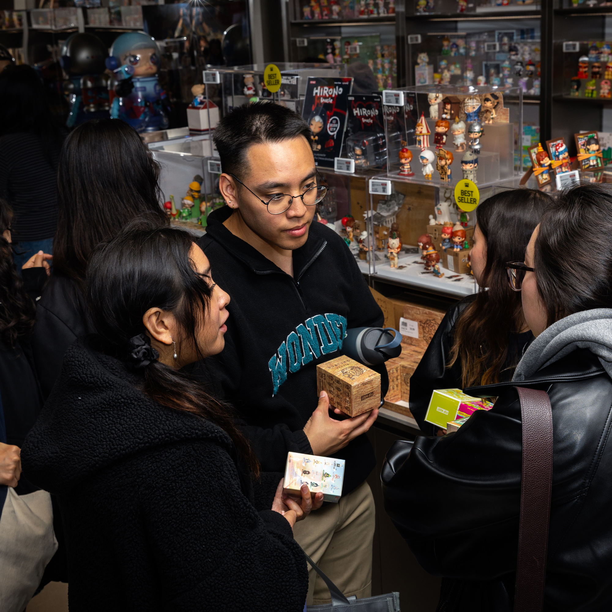 Several people are gathered in front of a glass display case with various collectibles and figurines. They are conversing and holding small boxed items.