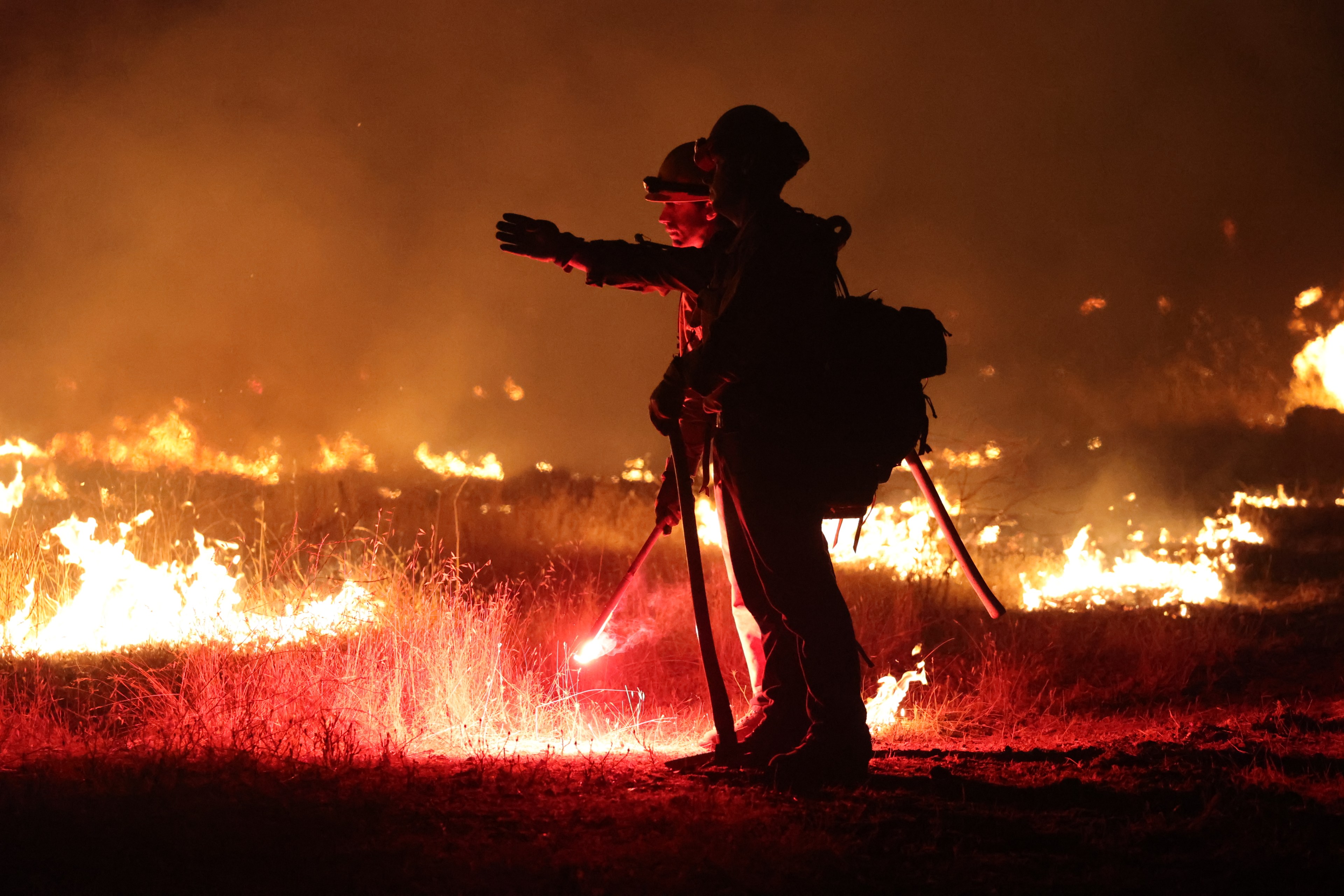 The silhouette of a firefighter is seen standing in the foreground as a grassy area burns behind him.