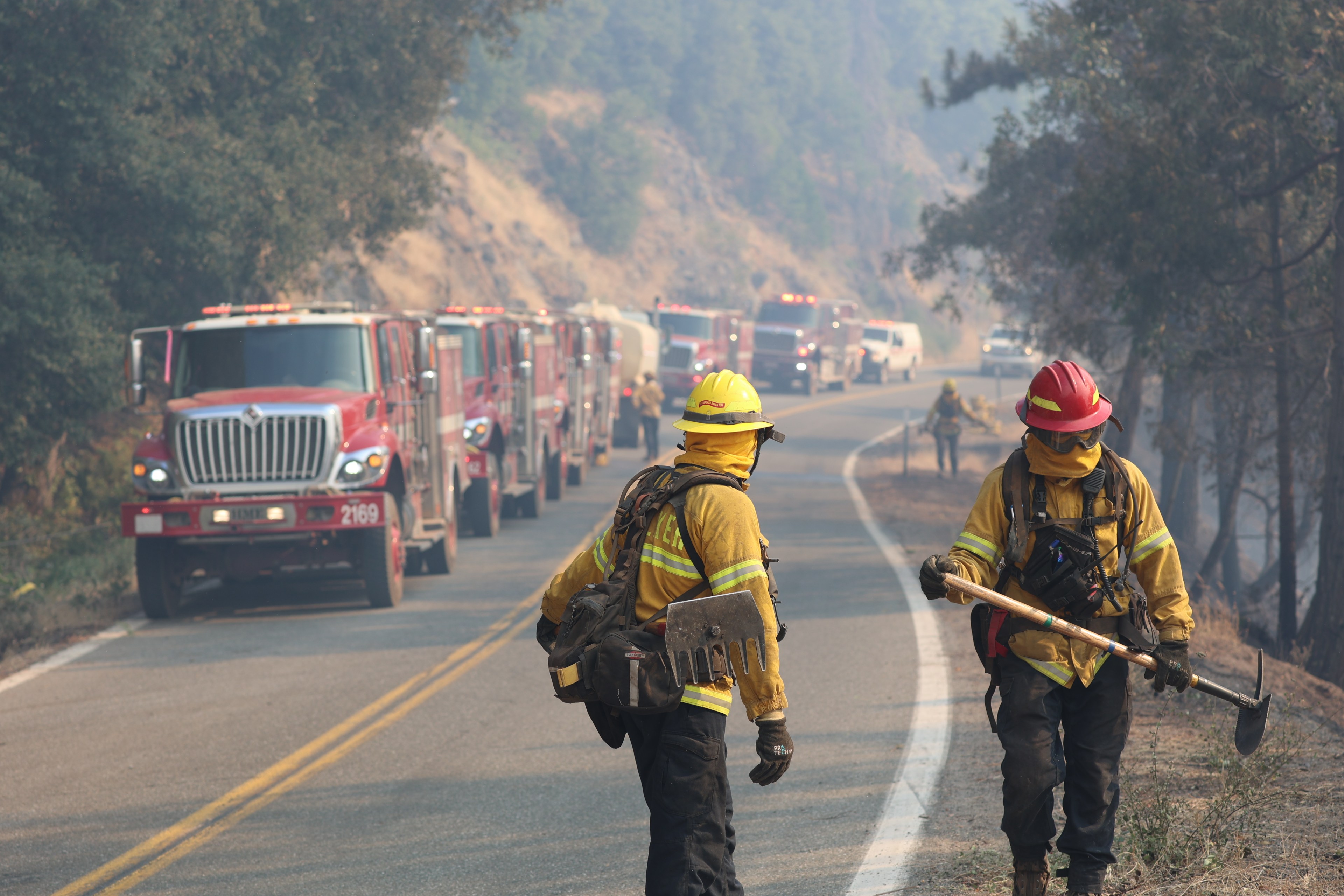 Firefighters stand along a road and several firetrucks are lined up near them.