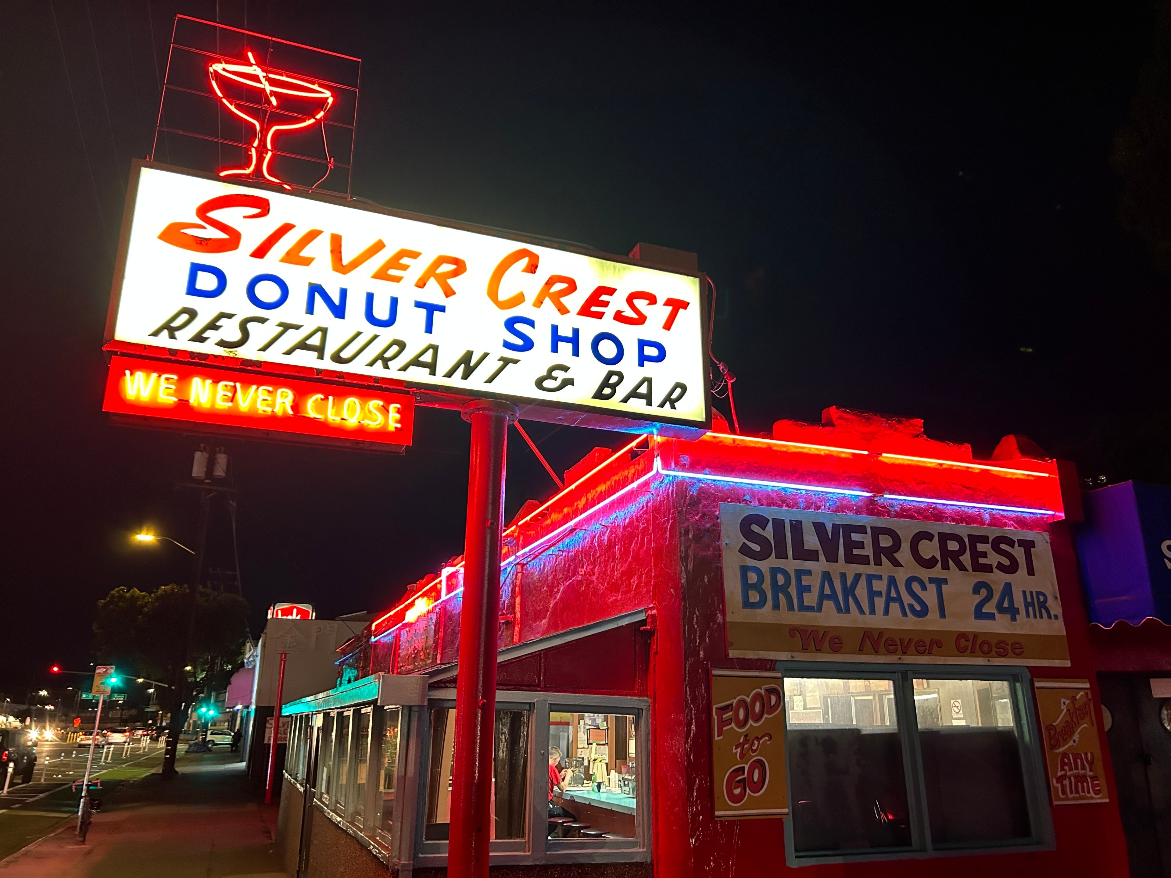 A neon-lit sign at night shows &quot;Silver Crest Donut Shop, Restaurant &amp; Bar, We Never Close,&quot; with other signage advertising 24-hour breakfast and food to go.