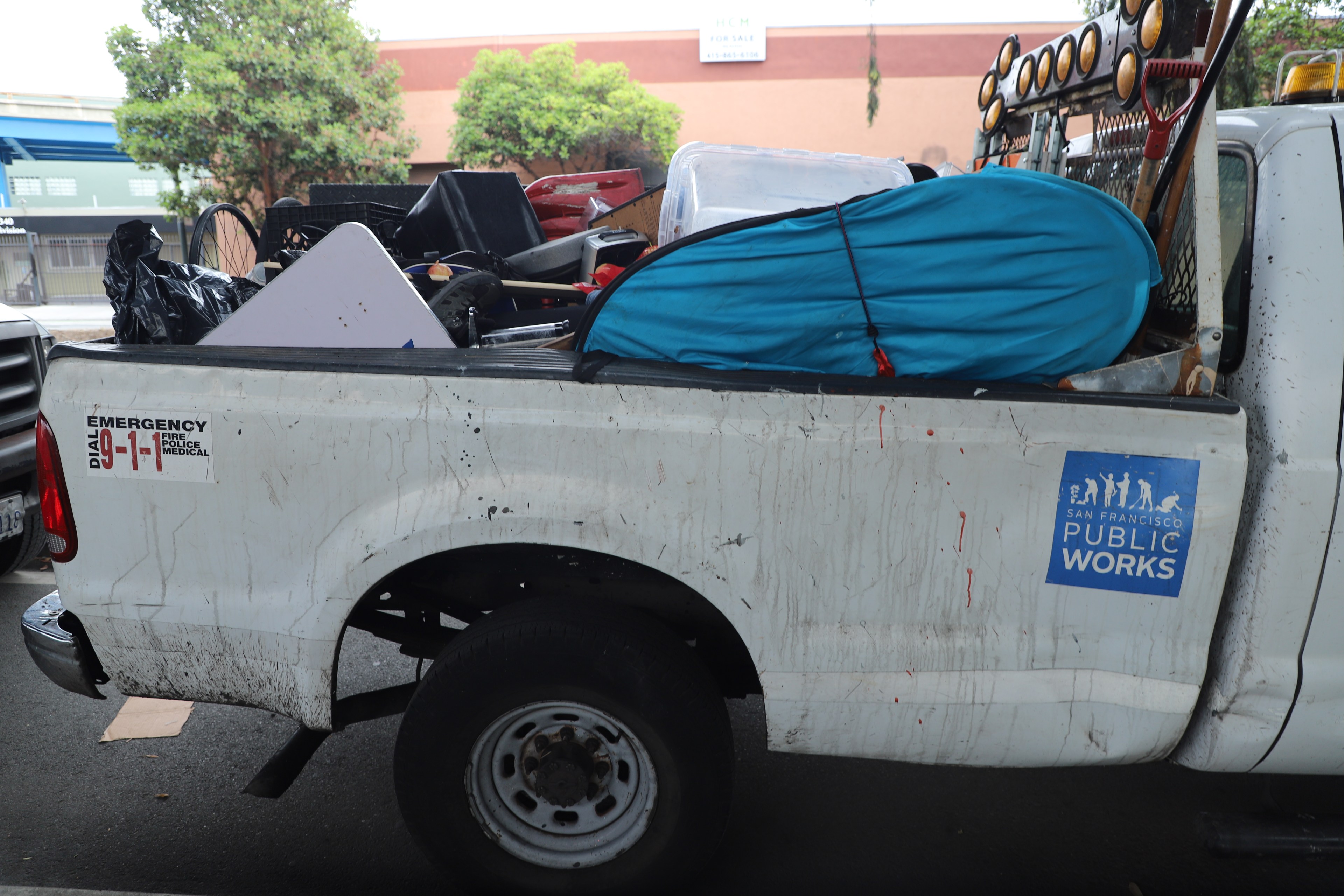 A San Francisco Public Works truck bed is filled with various items, including a blue tent, black bags, and other equipment. Trees and buildings are in the background.