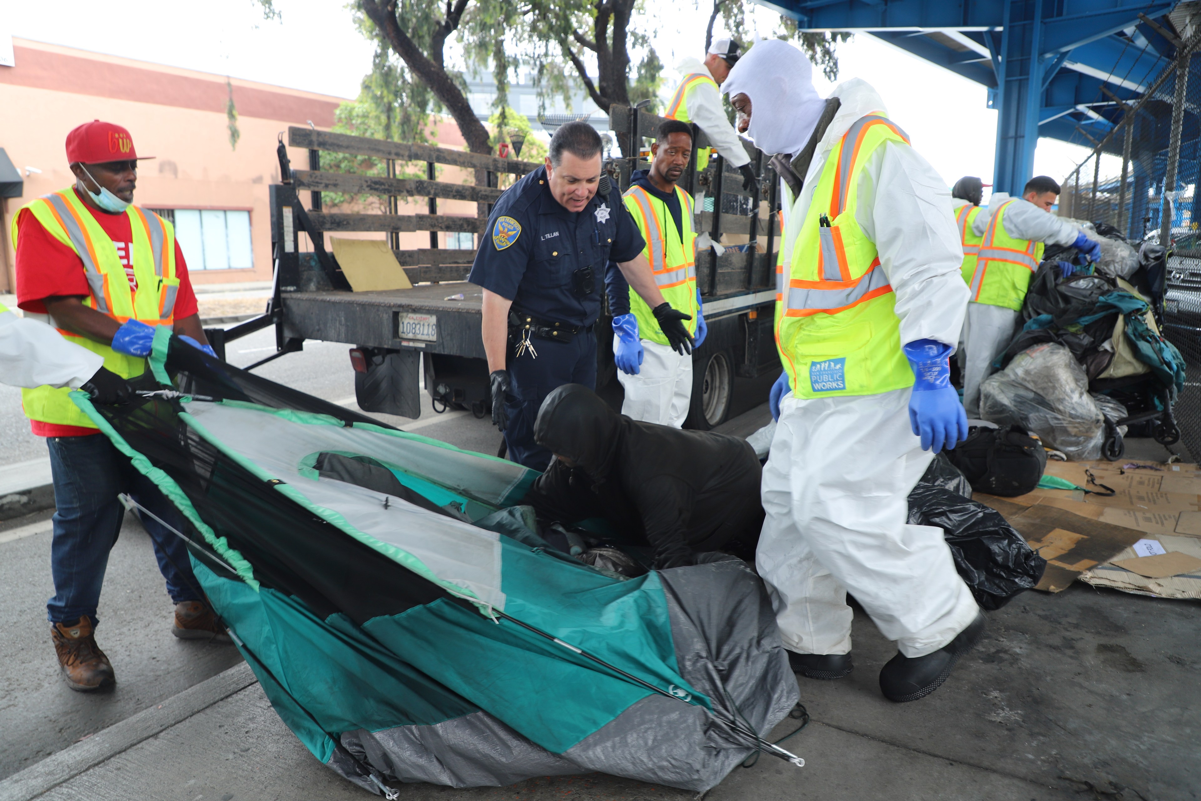 Several workers in protective suits and a police officer assist a person in dismantling a tent under a bridge, with a truck and other workers in the background.