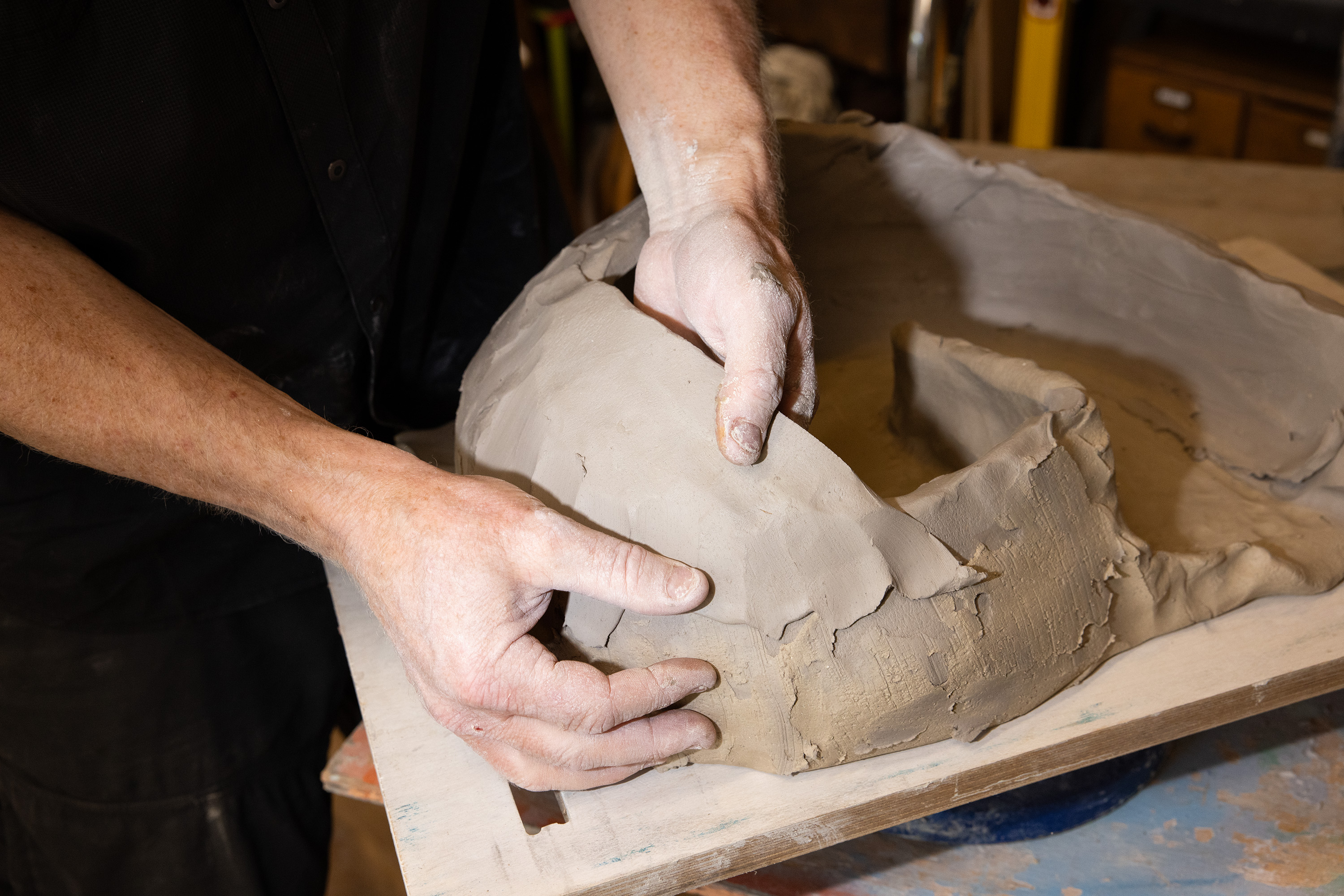 A person is working with a lump of clay, shaping and smoothing it with their hands, which are covered in clay residue. They are wearing a dark shirt.