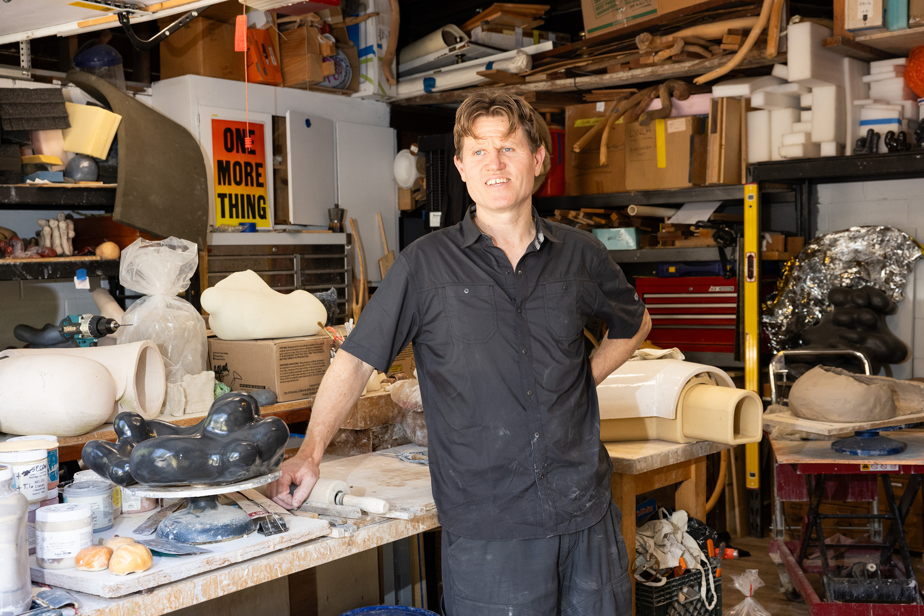 A man stands in a cluttered workshop filled with various art supplies and sculptures. He is wearing a black shirt and has one hand on his hip, smiling at the camera.