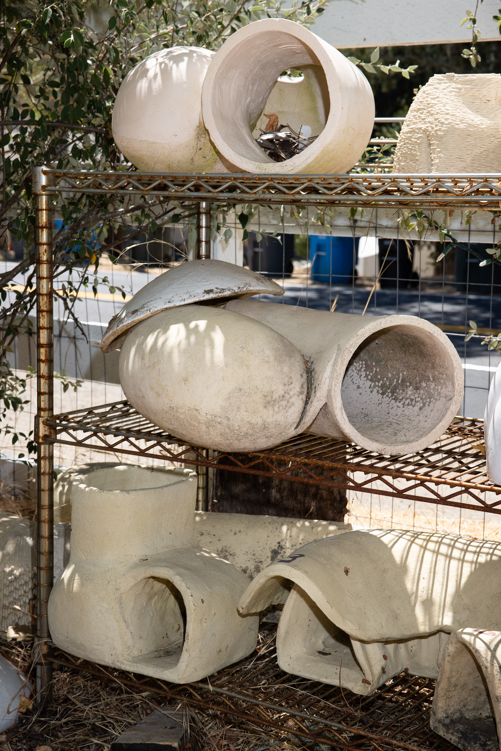 The image shows various white, cylindrical ceramic pieces stacked on metal shelves outdoors, with a few leaves and branches in the background.