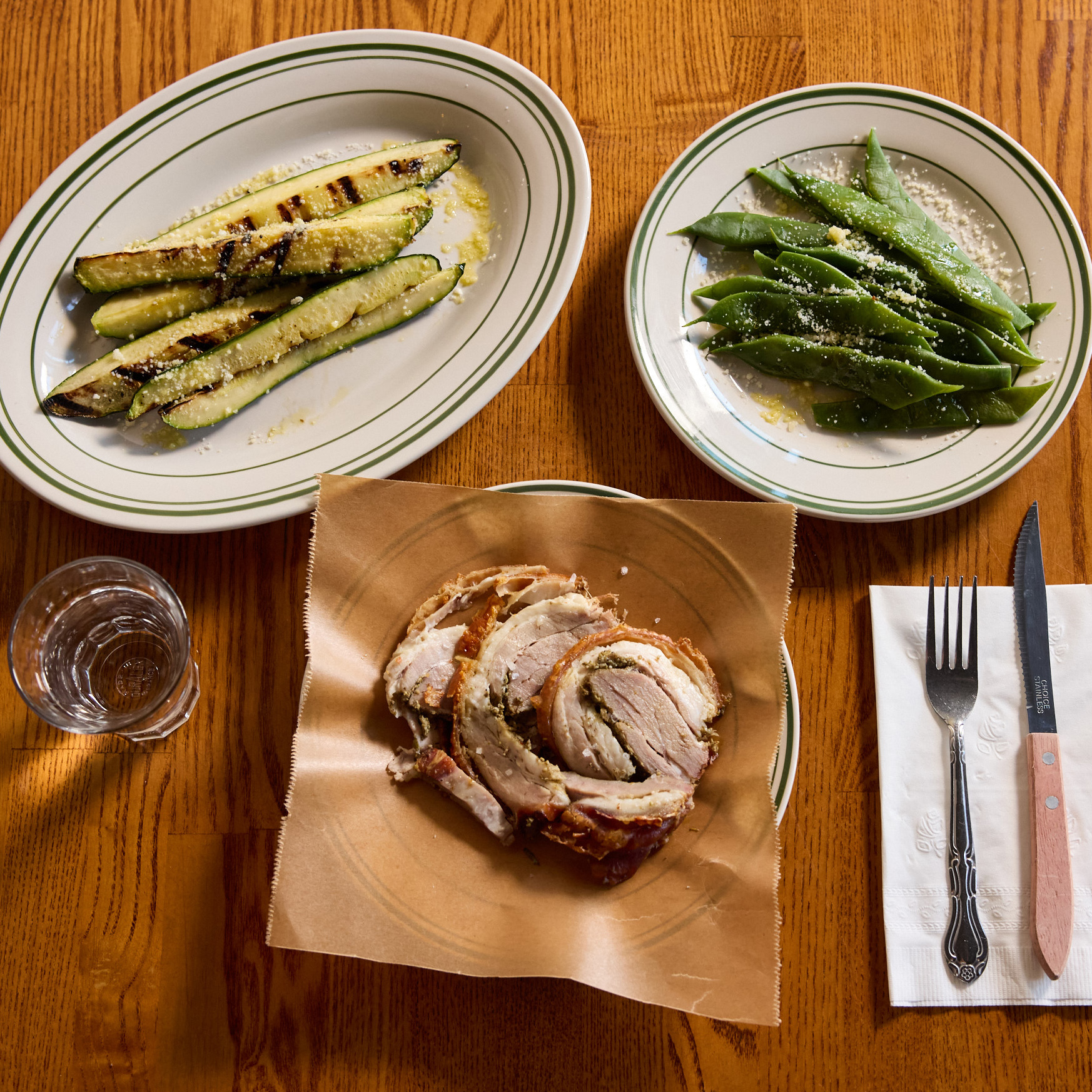 The image features plates of grilled zucchini, green beans, and sliced meat on a wooden table. A glass of water, fork, and knife are also present.