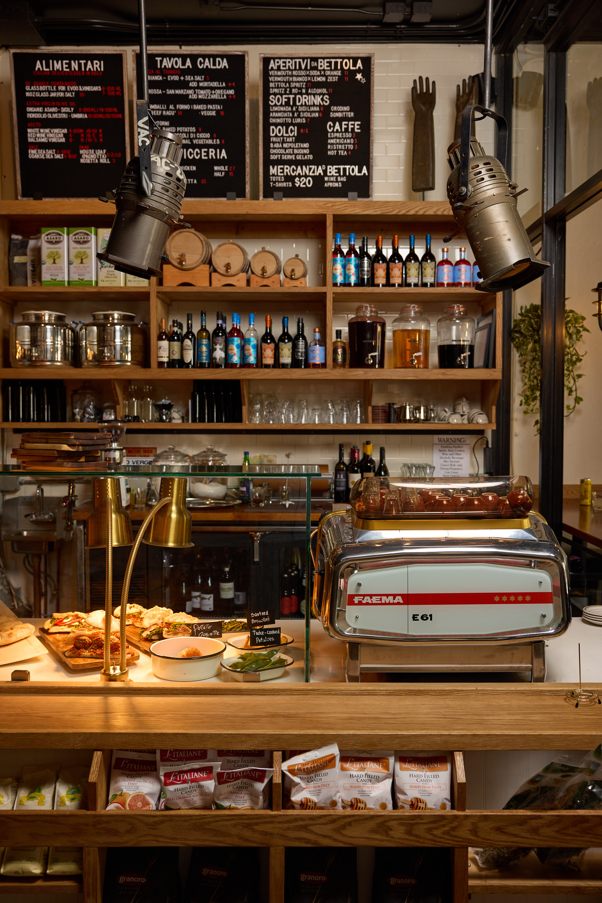 The image displays a cozy cafe with a counter showcasing various pastries, a coffee machine, wooden shelves with wine bottles, glasses, snacks, and a menu board above.