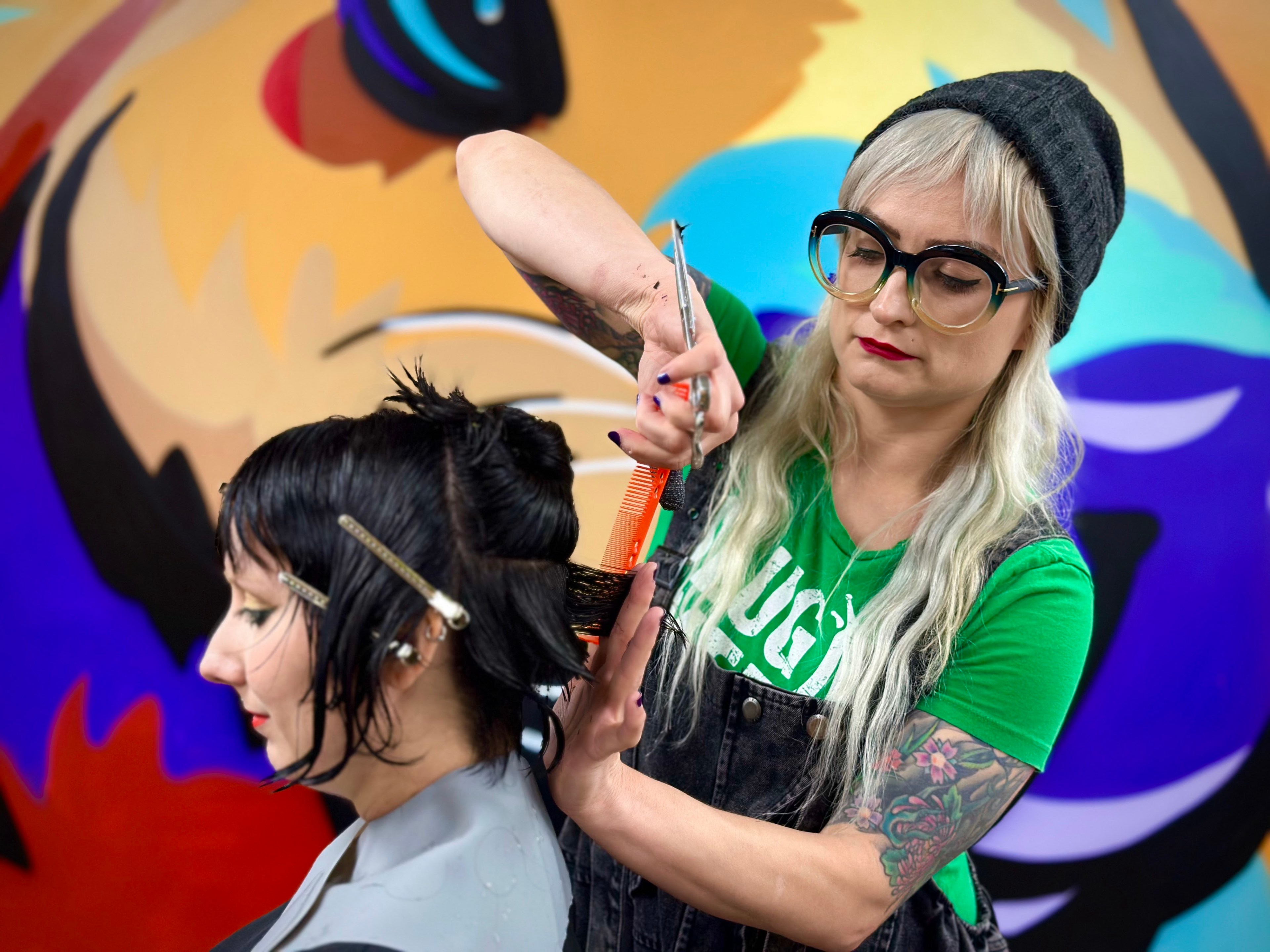 A hairstylist with long blonde hair, large glasses, a beanie, and tattoos cuts a woman's short black hair with scissors against a colorful, abstract mural backdrop.