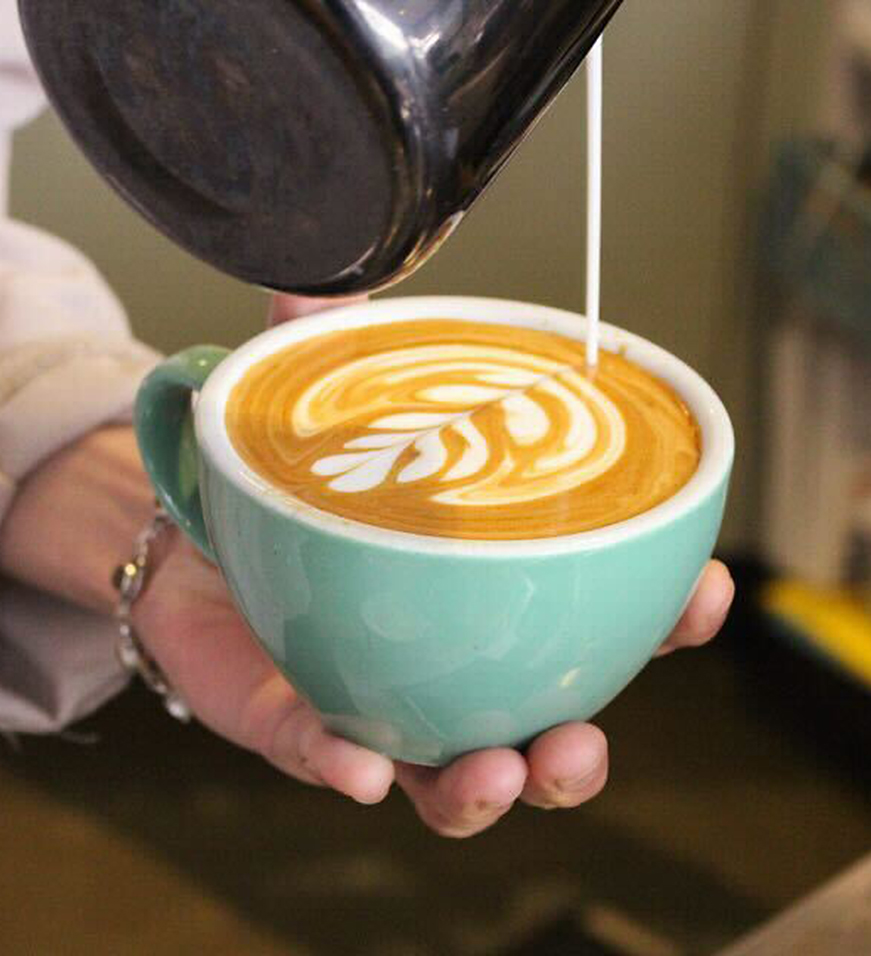 A hand holds a light blue mug of coffee, while milk is being poured from a metal pitcher to create a leaf-shaped latte art on the surface.