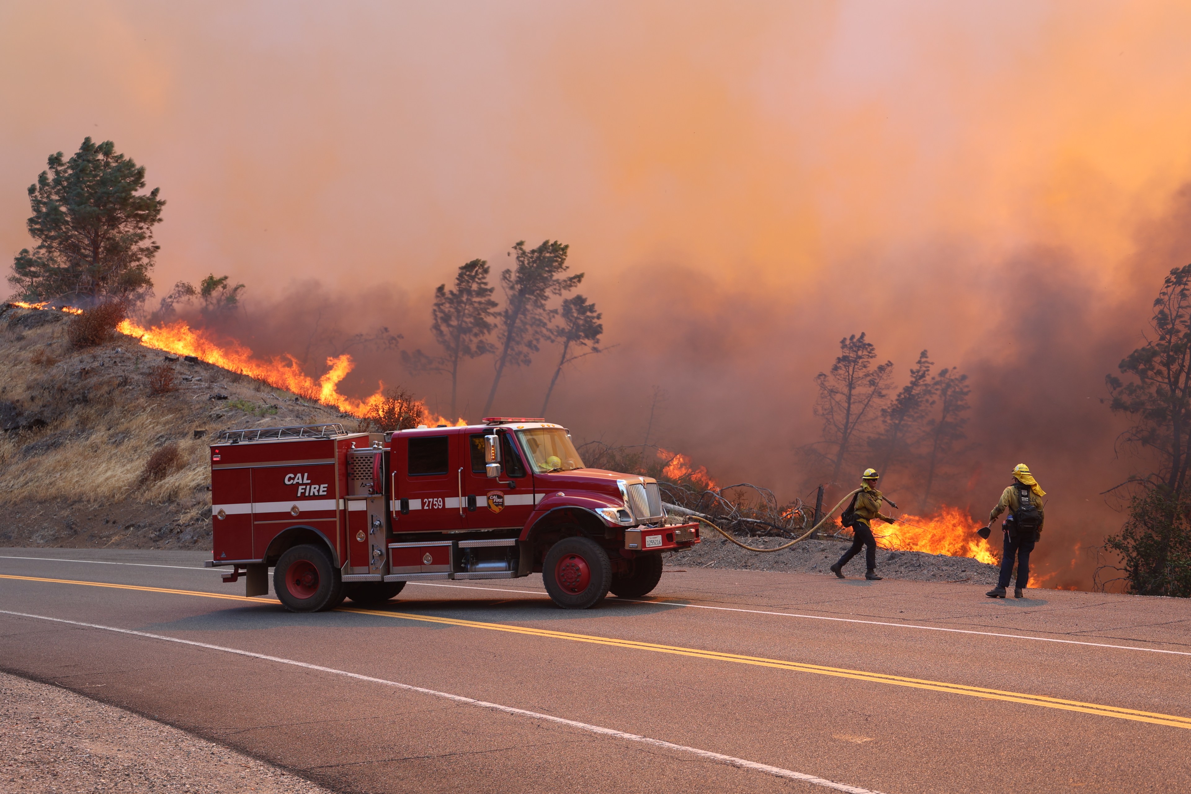 Two firefighters in yellow gear work to control a wildfire near a road. Flames blaze through dry brush, and a red fire truck is parked nearby.