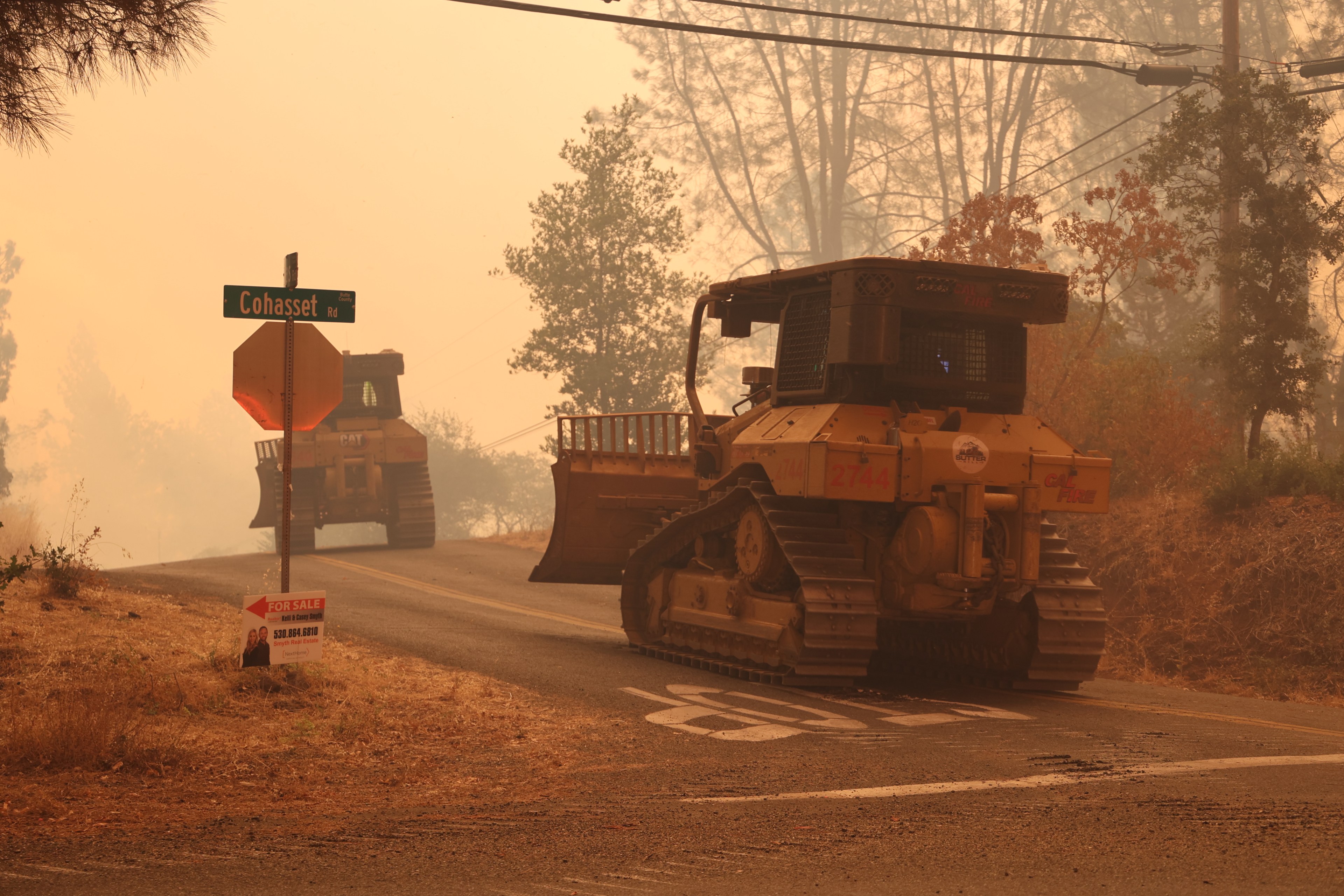 Two large bulldozers are on a smoky, rural road, near a signpost reading &quot;Cohasset Rd&quot; and a &quot;For Sale&quot; sign. The area is hazy, likely due to wildfire conditions.