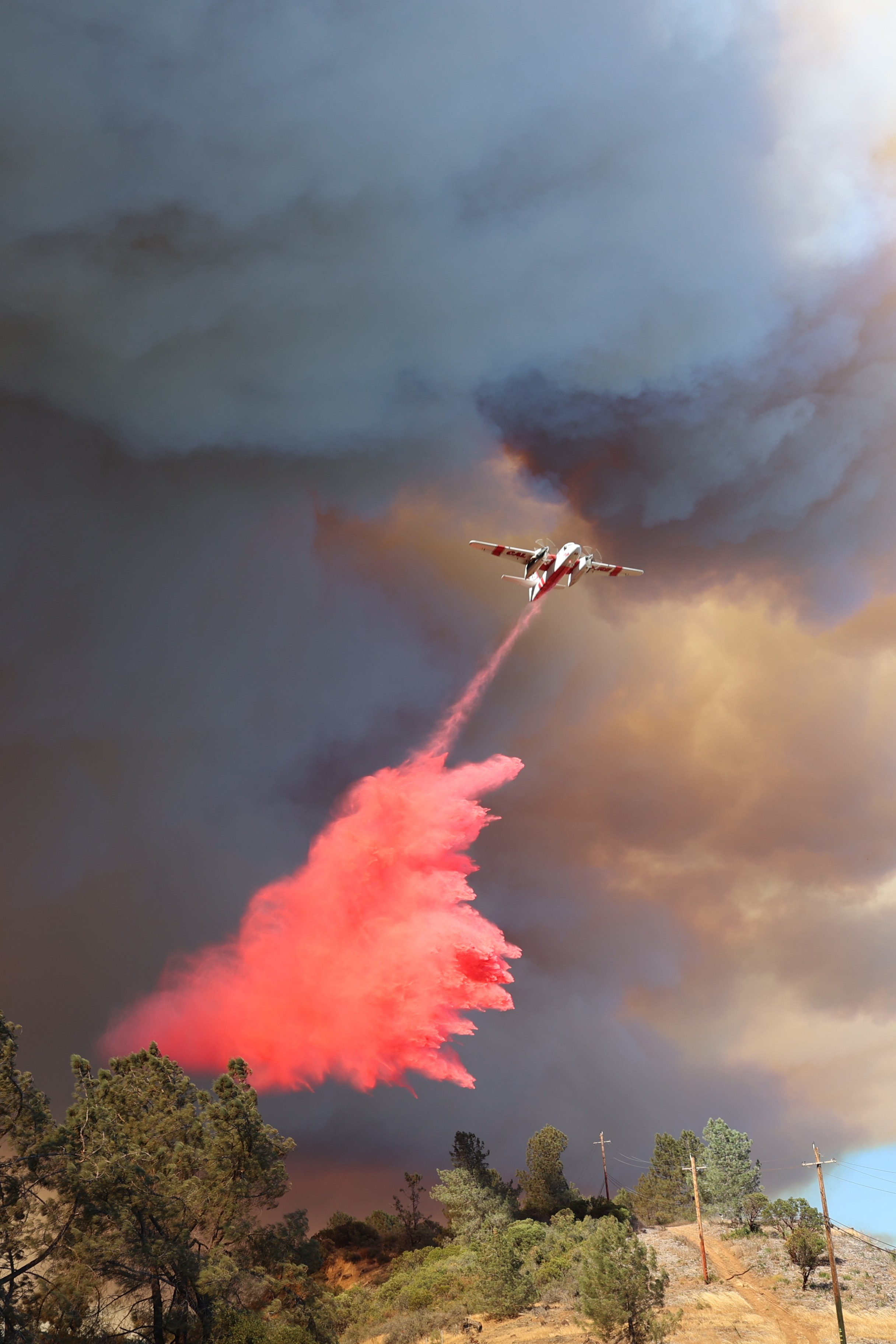 A plane releases red fire retardant over a wildfire. Huge clouds of smoke fill the sky, and trees along with a hilly landscape are visible below.