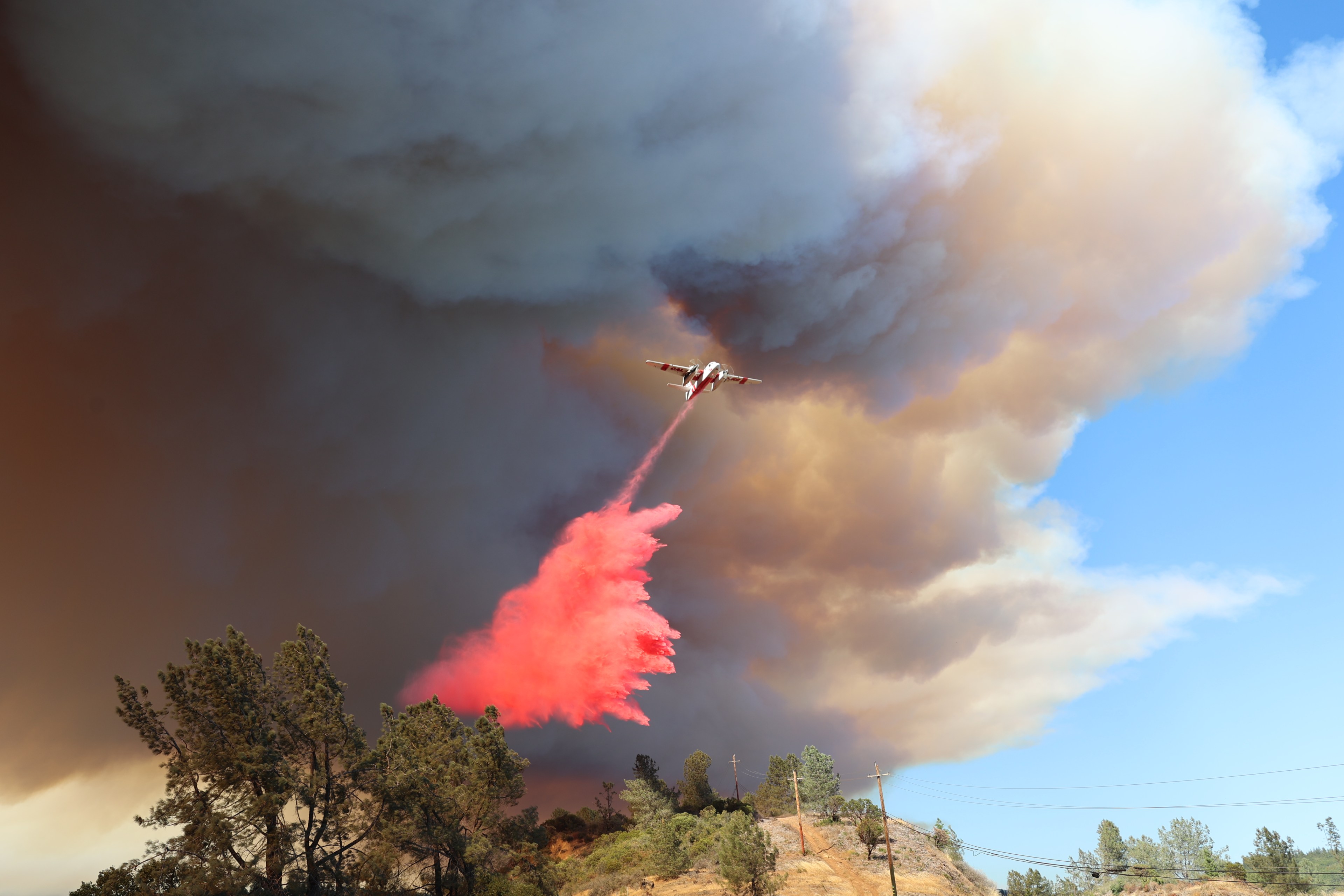 A plane releases red fire retardant over a wildfire. Huge clouds of smoke fill the sky, and trees along with a hilly landscape are visible below.