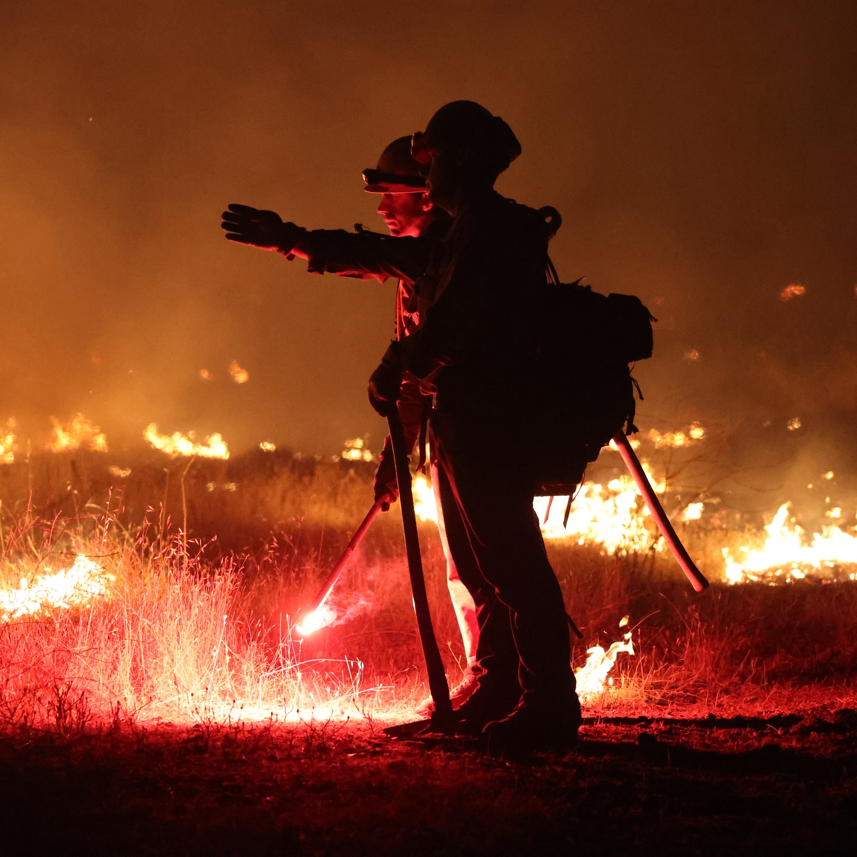 Two firefighters in protective gear are battling a wildfire at night, surrounded by flames and smoke. One is pointing while the other holds a glowing flare.