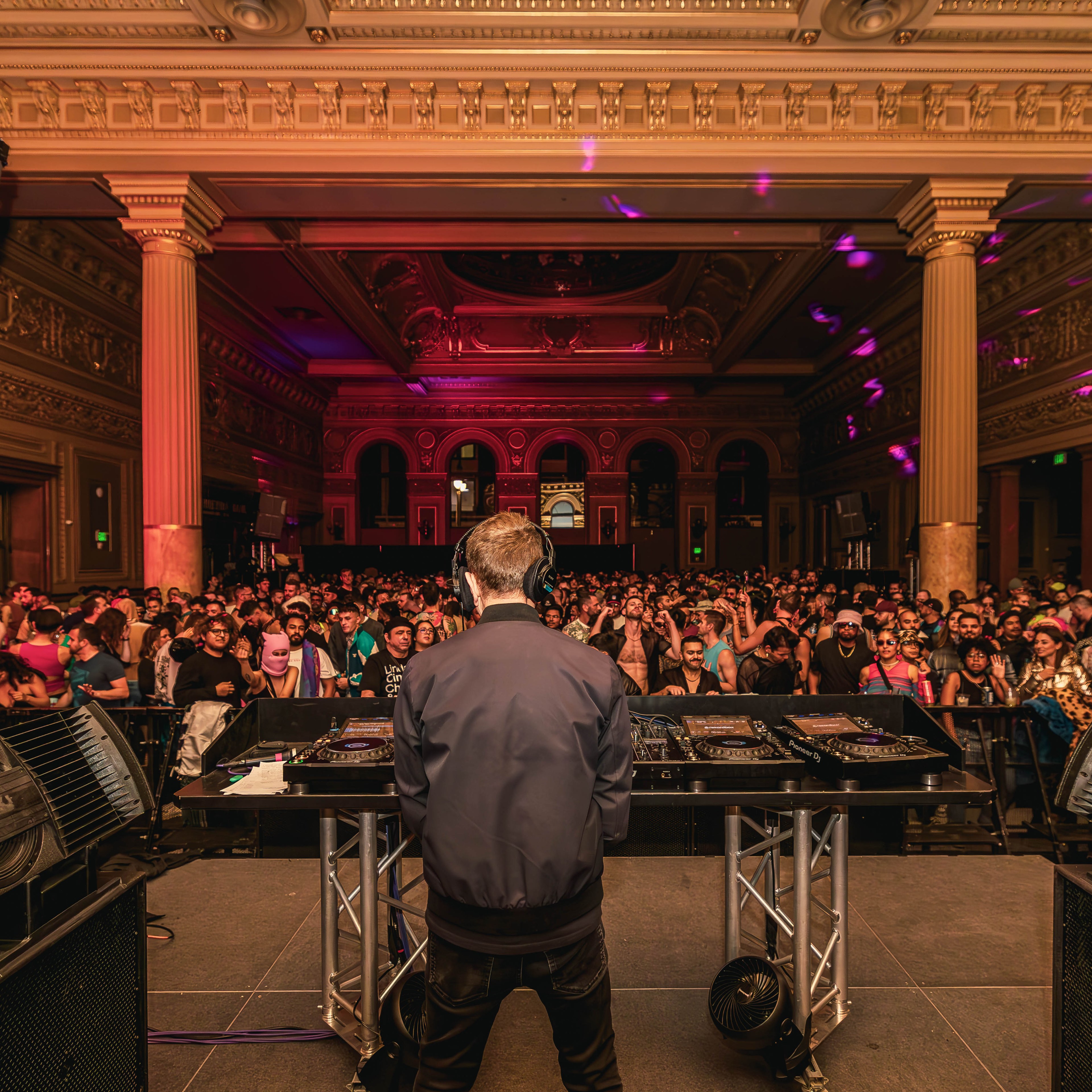 A DJ, seen from behind, performs on stage with large speakers in an opulent hall filled with a vibrant crowd, under colorful lighting, inside a grand, ornate building.