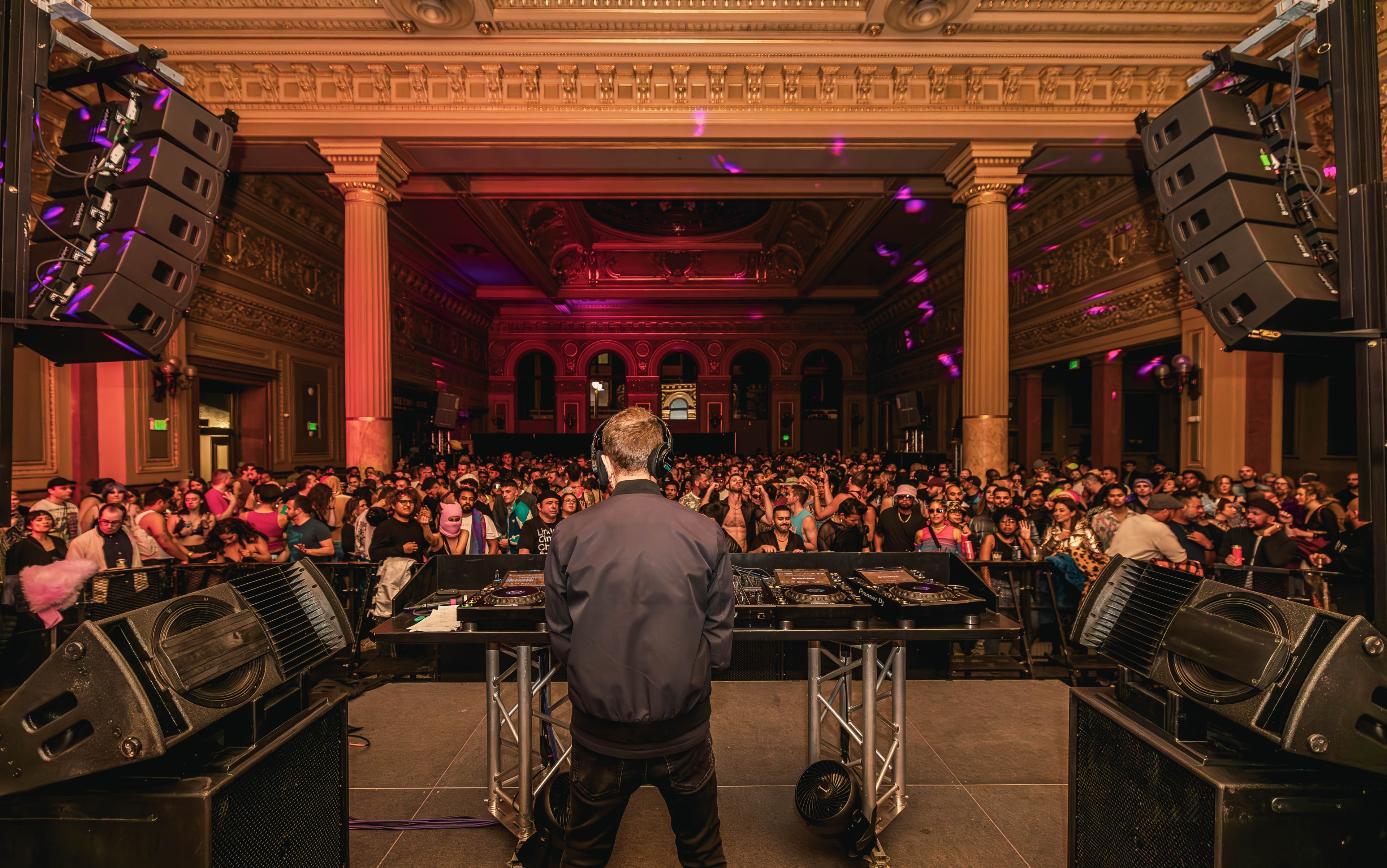 A DJ, seen from behind, performs on stage with large speakers in an opulent hall filled with a vibrant crowd, under colorful lighting, inside a grand, ornate building.