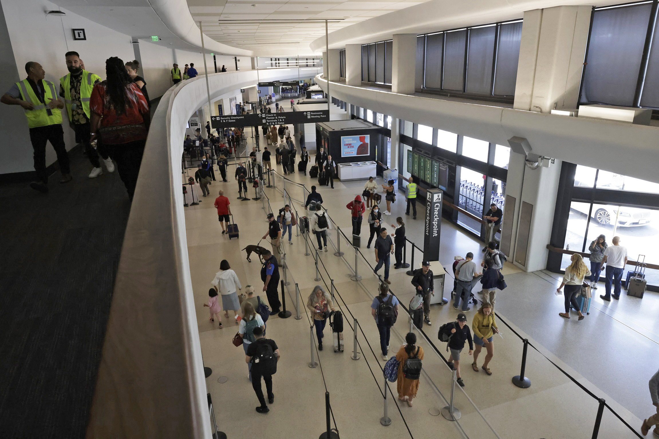 A busy airport terminal shows travelers lined up at security checkpoints with airport staff nearby. Large windows on the right let in natural light.