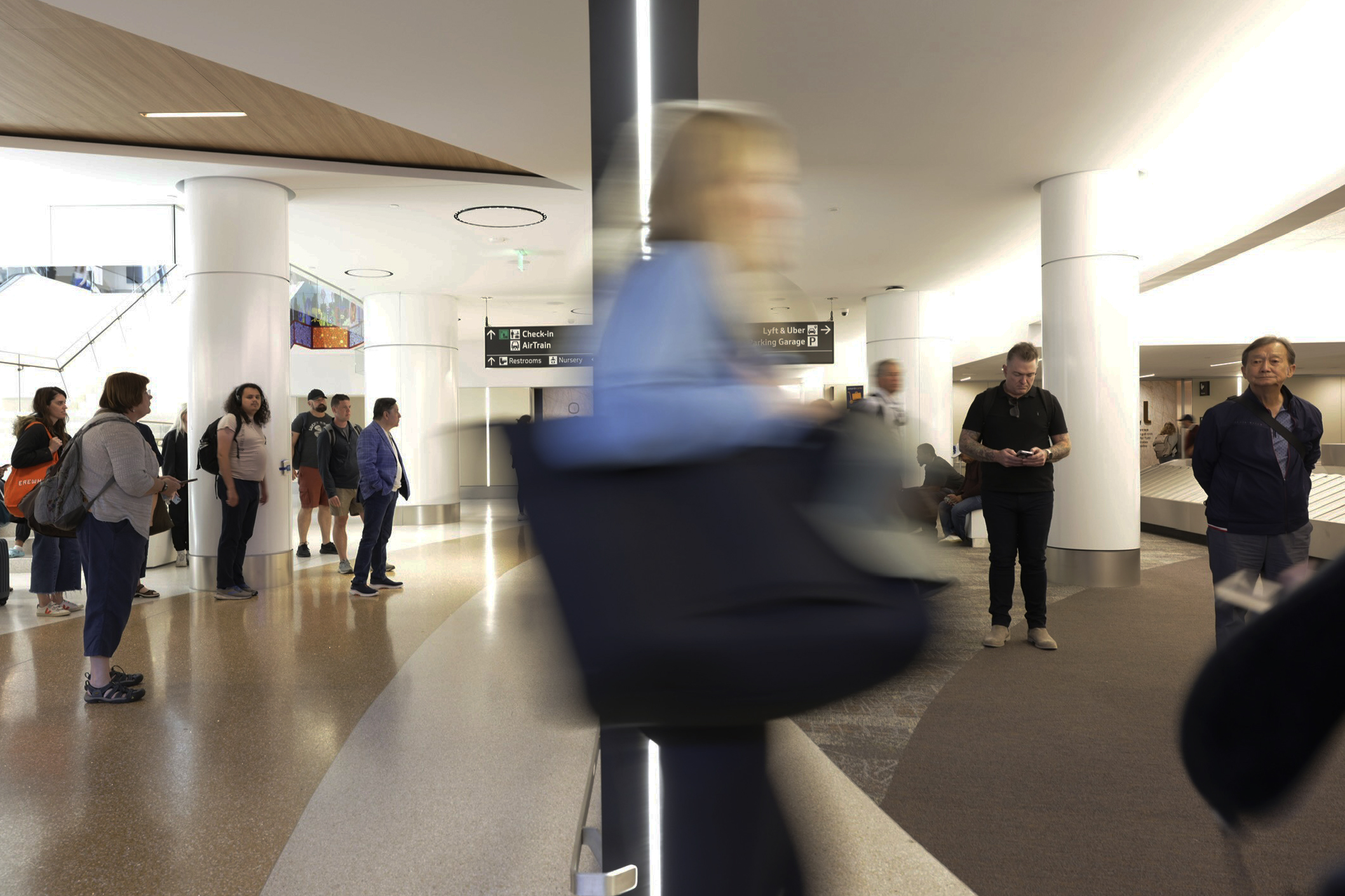 The image shows people standing in an airport terminal, with some waiting and one person blurred in motion. Signage for check-in, AirTrain, and restrooms is visible.