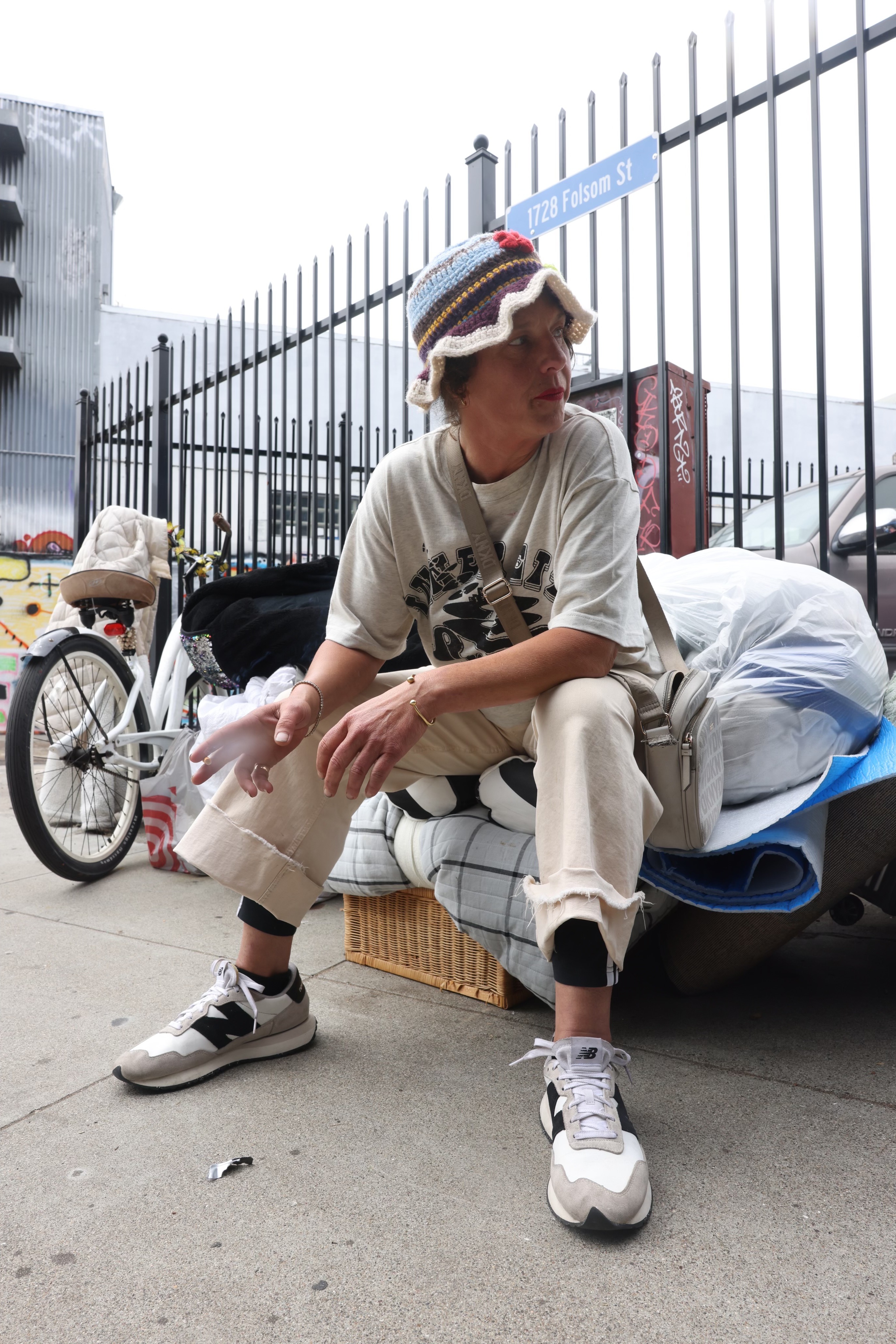 A person in a striped hat and casual clothing sits on a makeshift bench with belongings, next to a bike, near a fence labeled &quot;1728 Folsom St.&quot;