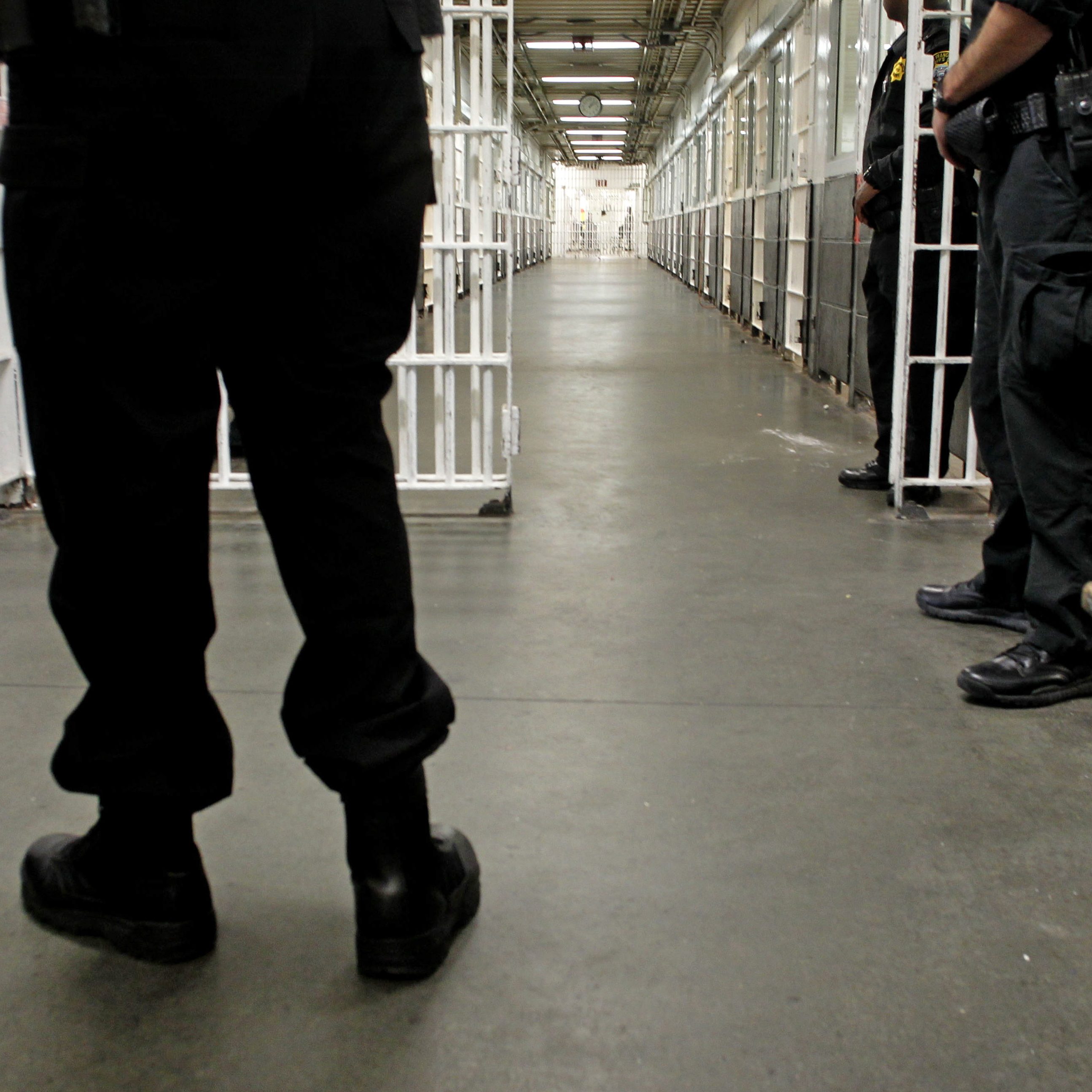 The image shows a corridor within a prison, lined with cells on both sides and guarded by officers in black uniforms. The ground is concrete, and the lighting is fluorescent.