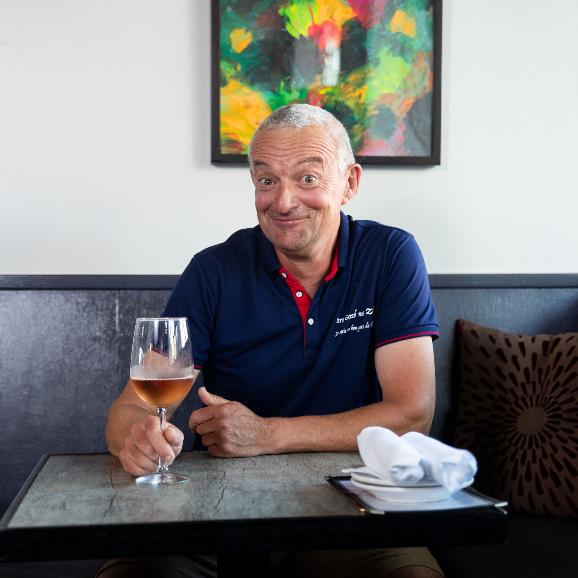 A man with short gray hair, wearing a navy polo shirt, sits at a table holding a glass of rosé wine. Behind him is a colorful abstract painting.