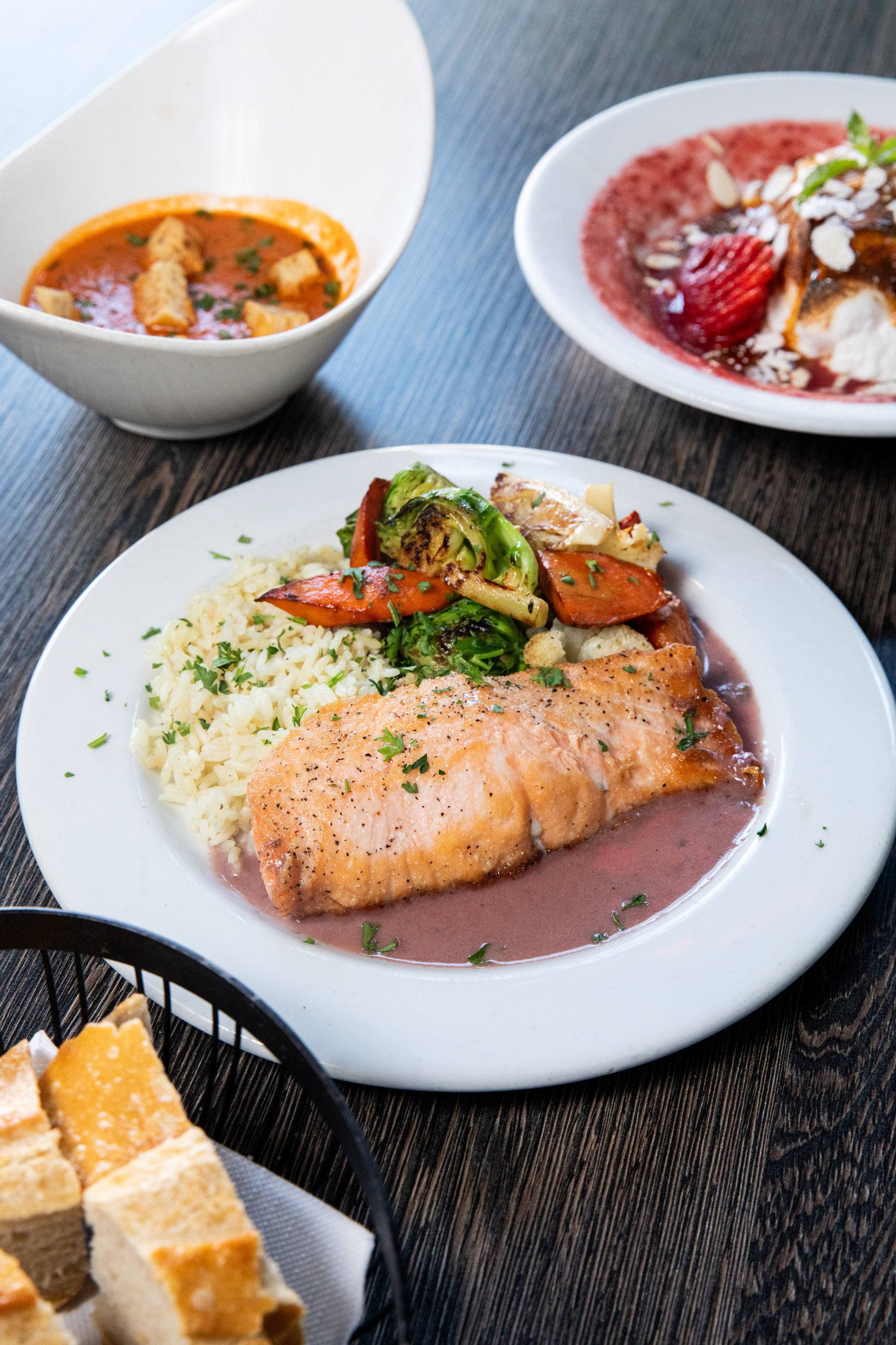 The image shows a plate with grilled salmon, rice, and mixed vegetables in a purple sauce. Nearby, there are bowls of soup, dessert with strawberries, and a basket of bread.