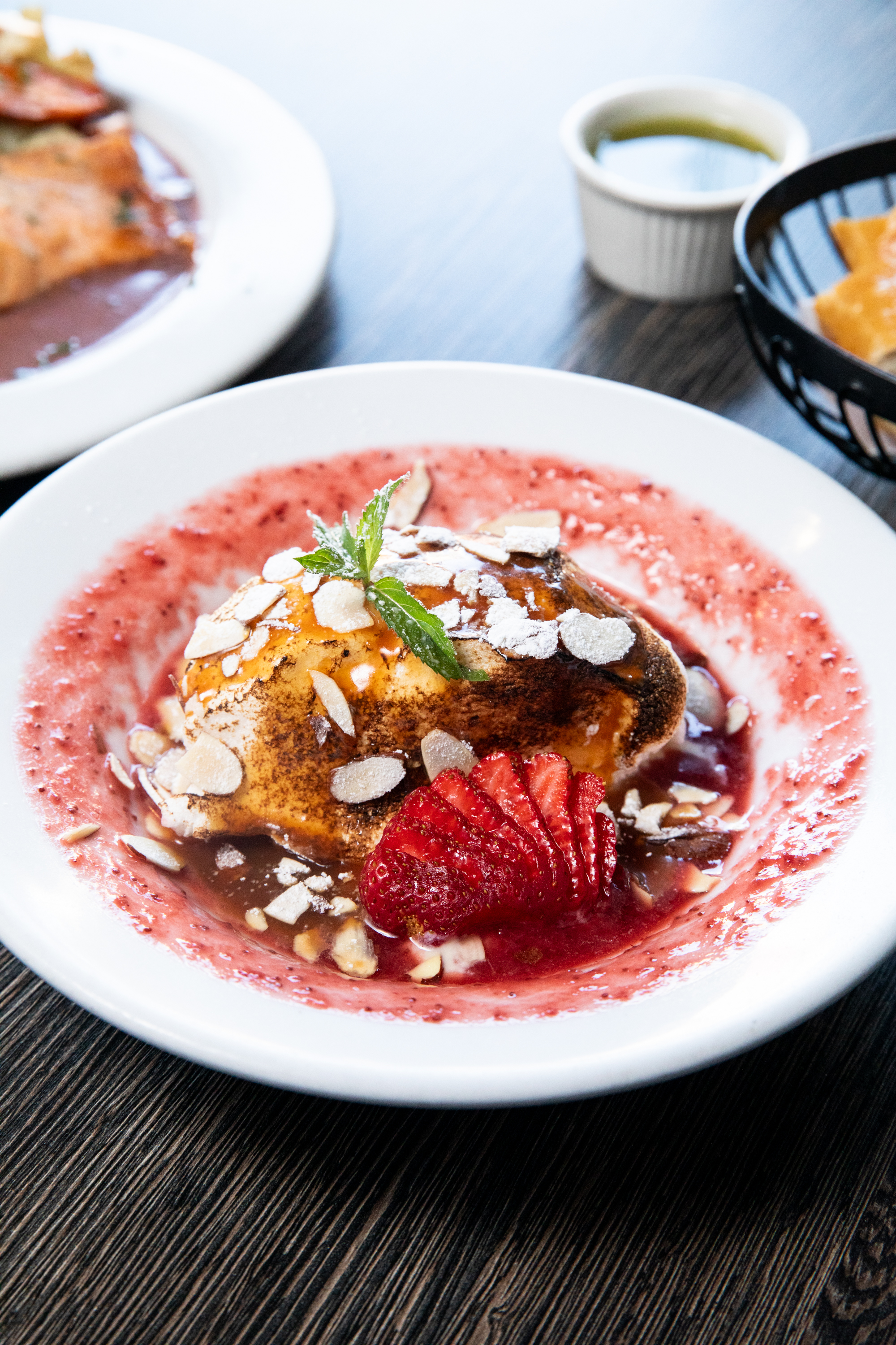 The image shows a dessert on a white plate with a dollop of cream, garnished with sliced strawberries, almond slivers, mint, and a berry sauce. Other dishes are blurred in the background.