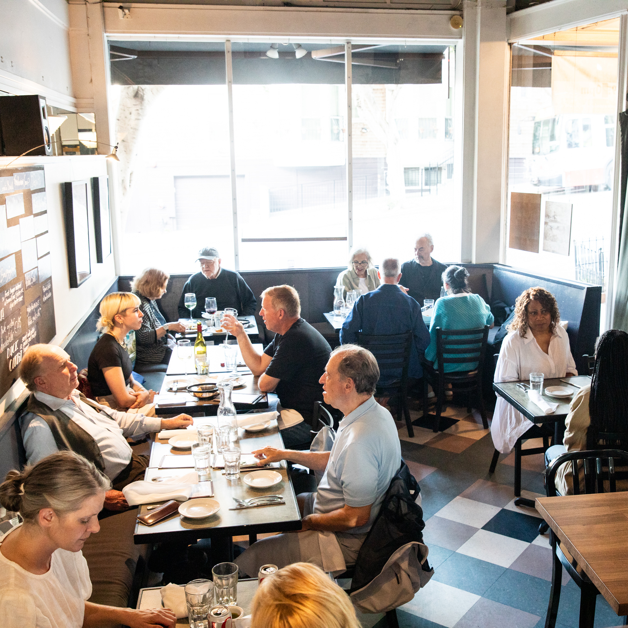 A cozy, brightly-lit restaurant features several groups of people seated at tables, engaged in conversation and dining. The room has large windows and simple decor.