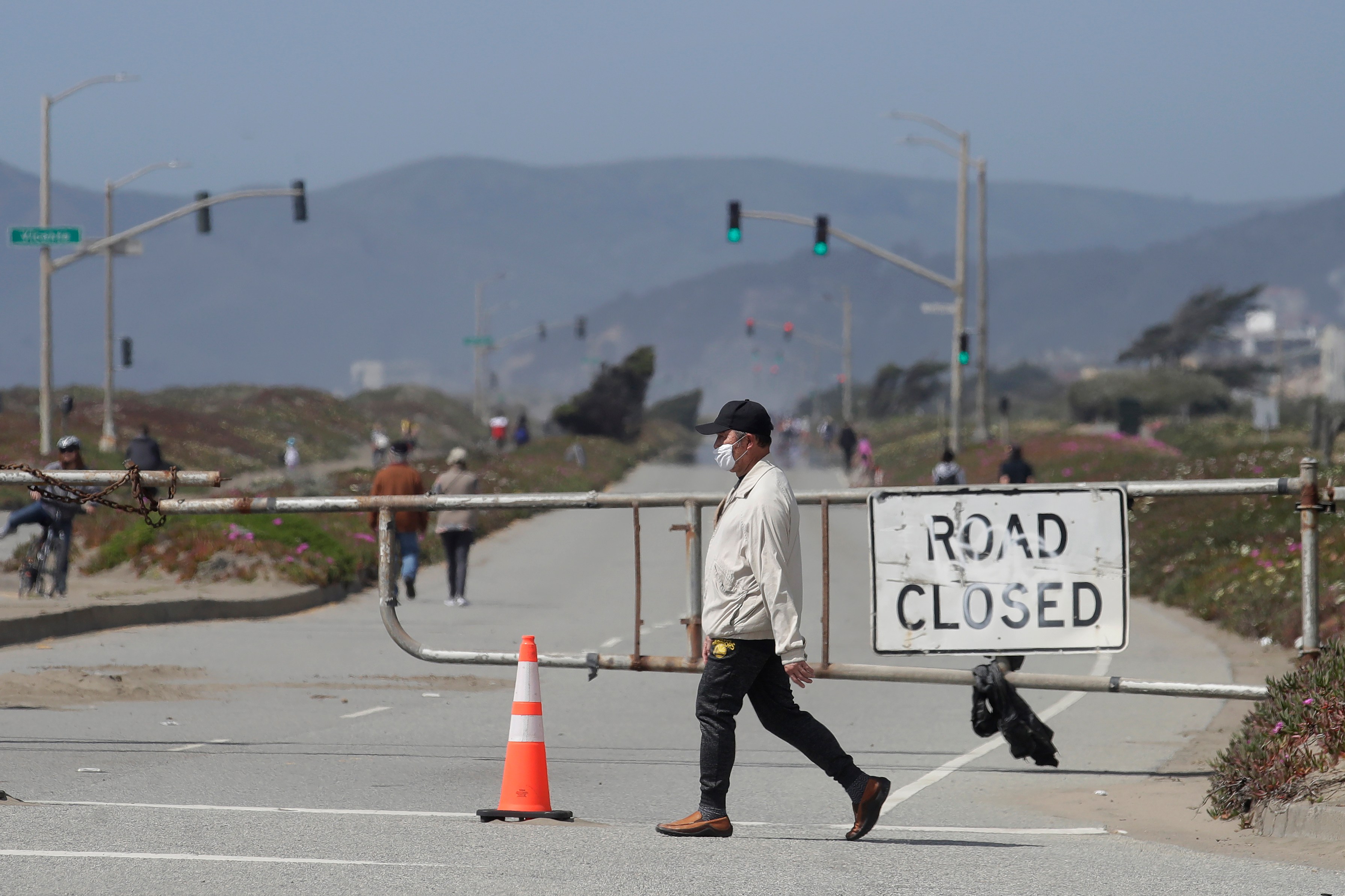 A man wearing a mask walks across a street closed off by a gate and a "Road Closed" sign. In the background, people are walking, and mountains are visible.