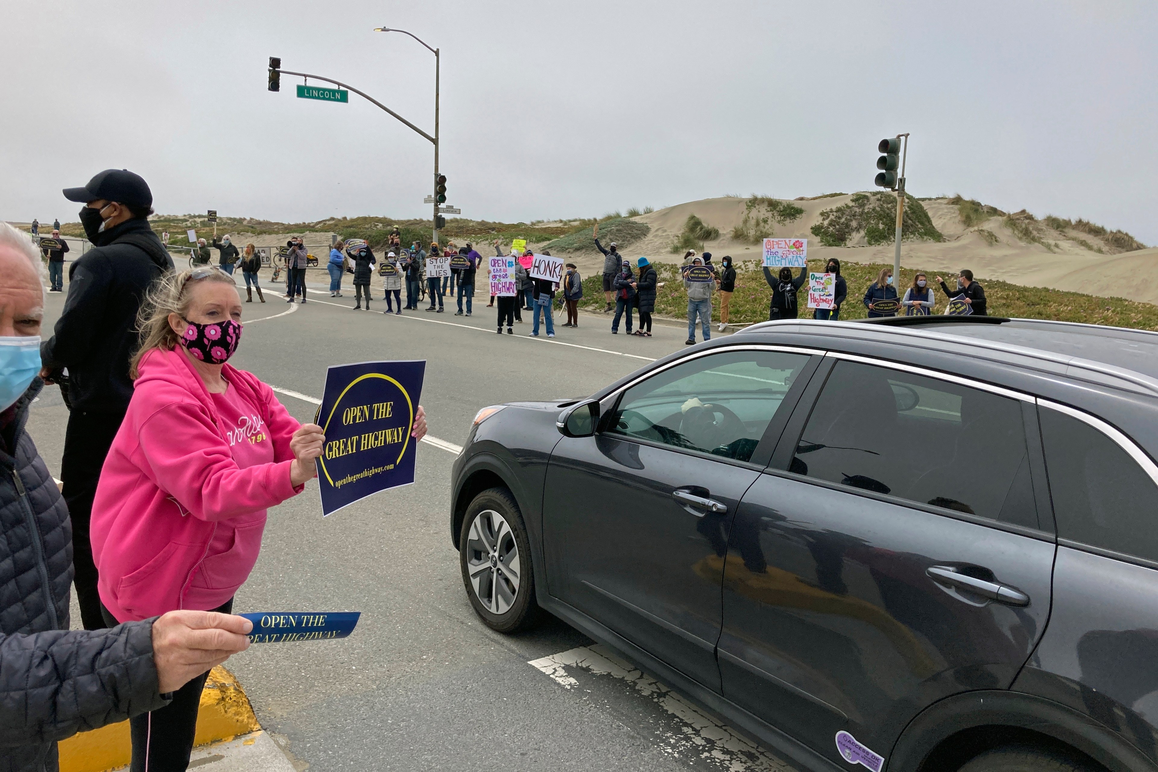 People are gathered at an intersection with signs protesting to &quot;Open the Great Highway,&quot; some standing on the roadside and others in a line across the street.