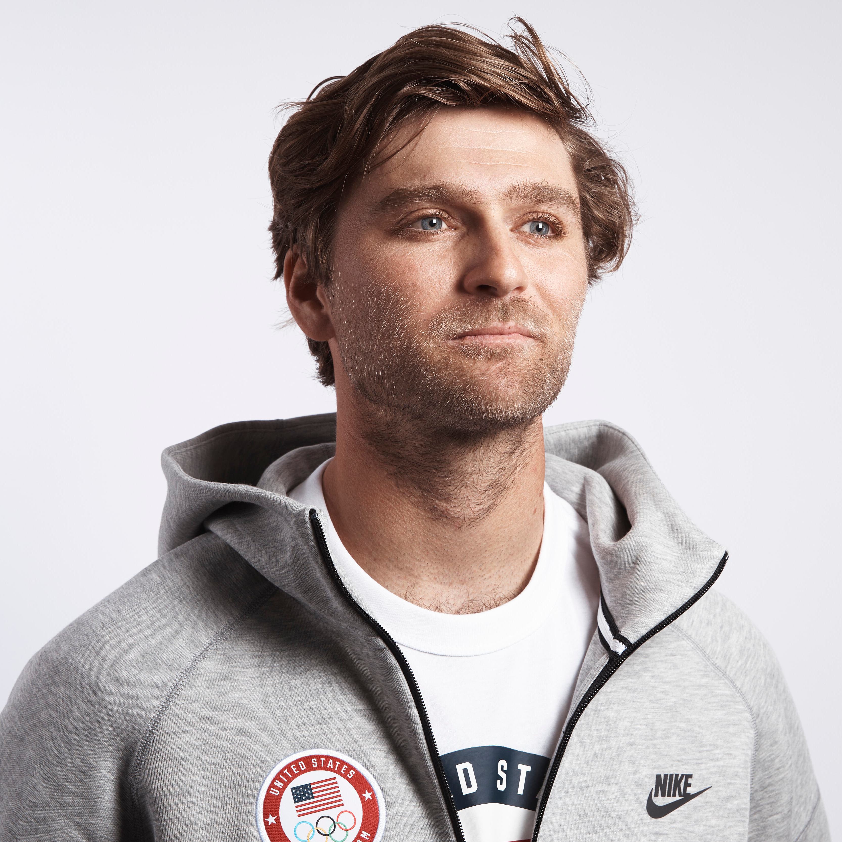 The image shows a man with light brown hair wearing a gray Nike hoodie with a United States Olympic logo patch and a white T-shirt underneath, looking slightly to his left.