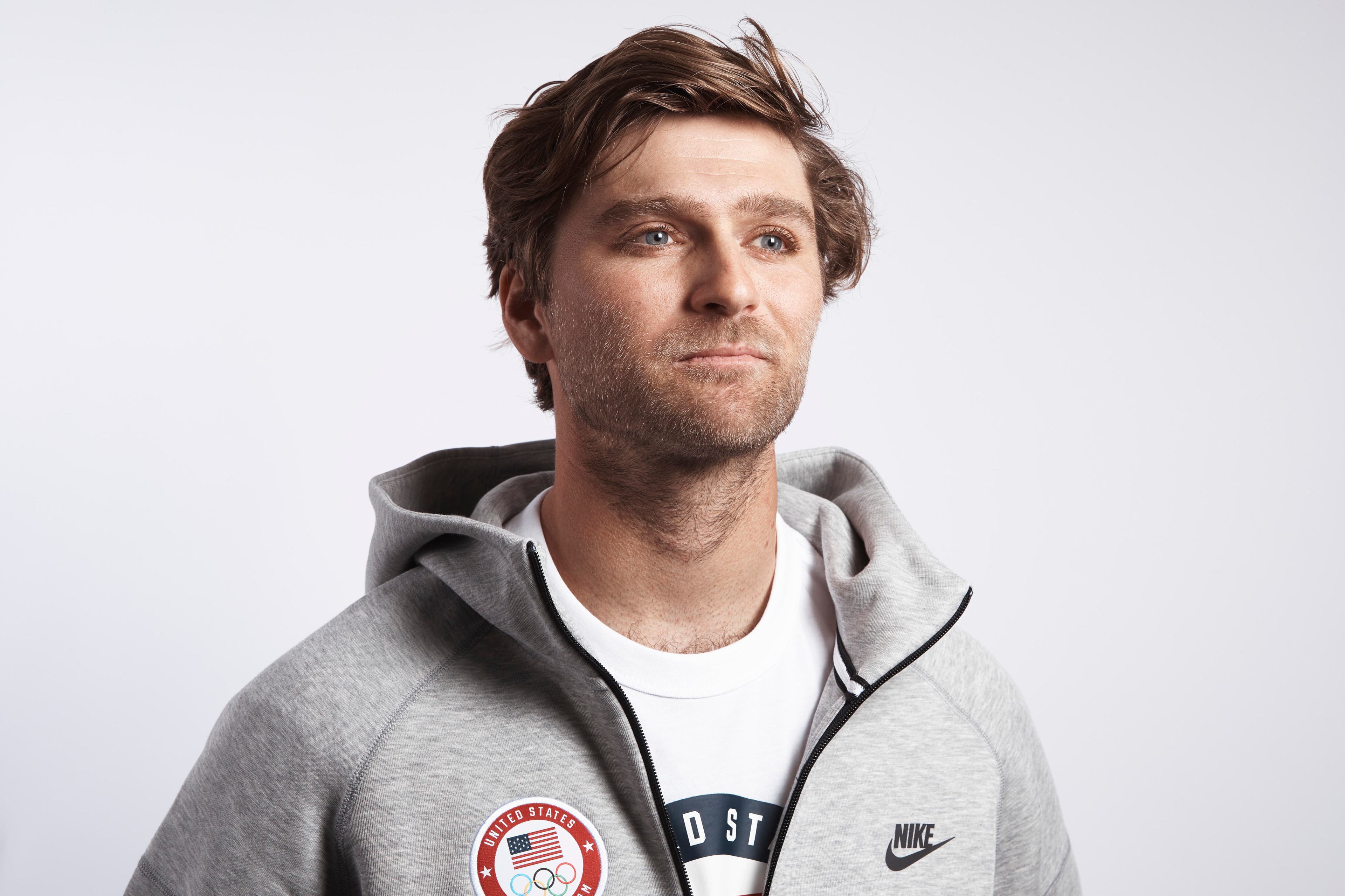 The image shows a man with light brown hair wearing a gray Nike hoodie with a United States Olympic logo patch and a white T-shirt underneath, looking slightly to his left.