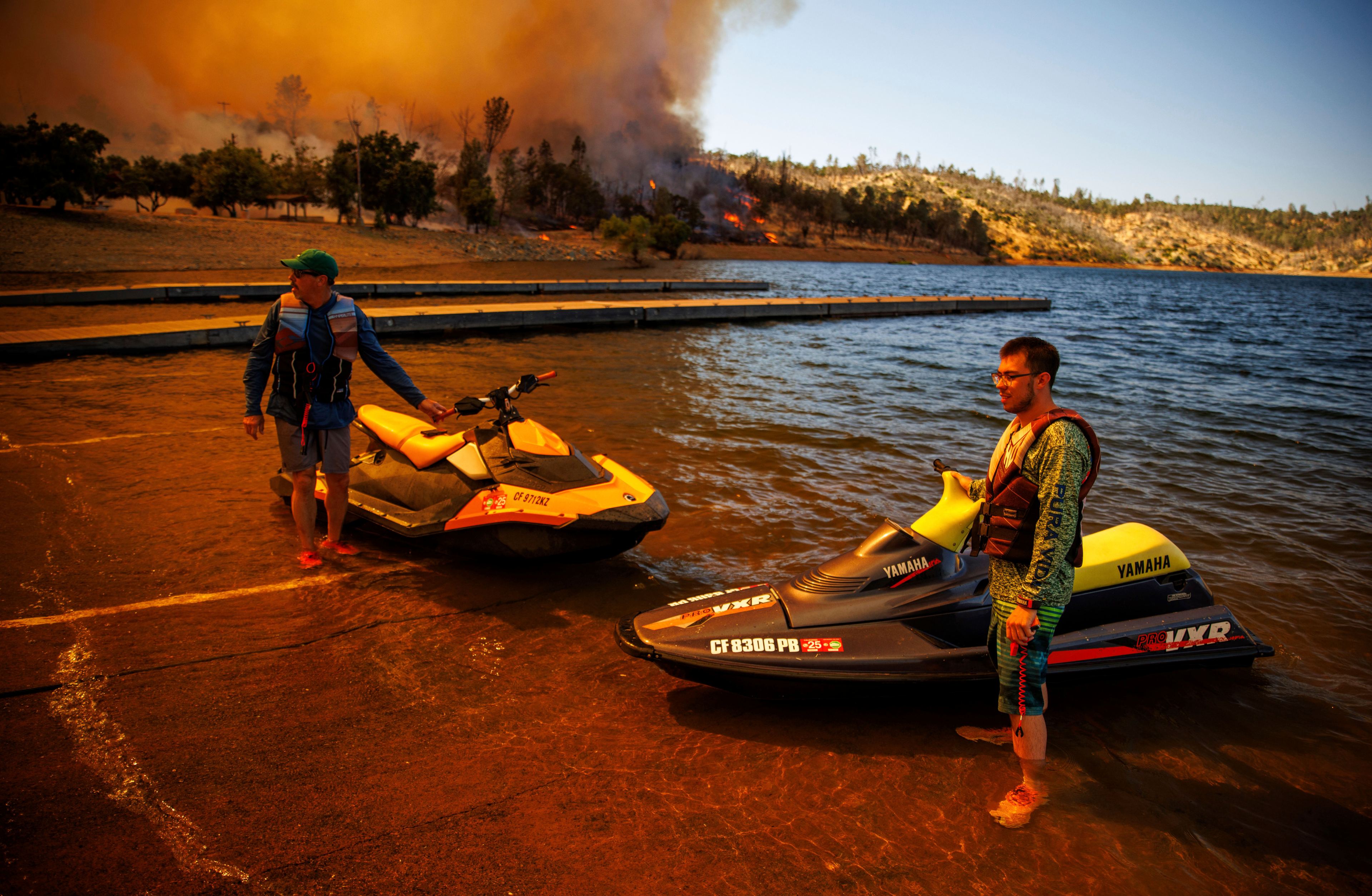 Two men wearing life vests are standing in shallow lake waters next to jet skis. Behind them, a forest fire burns intensely, filling the sky with thick smoke.