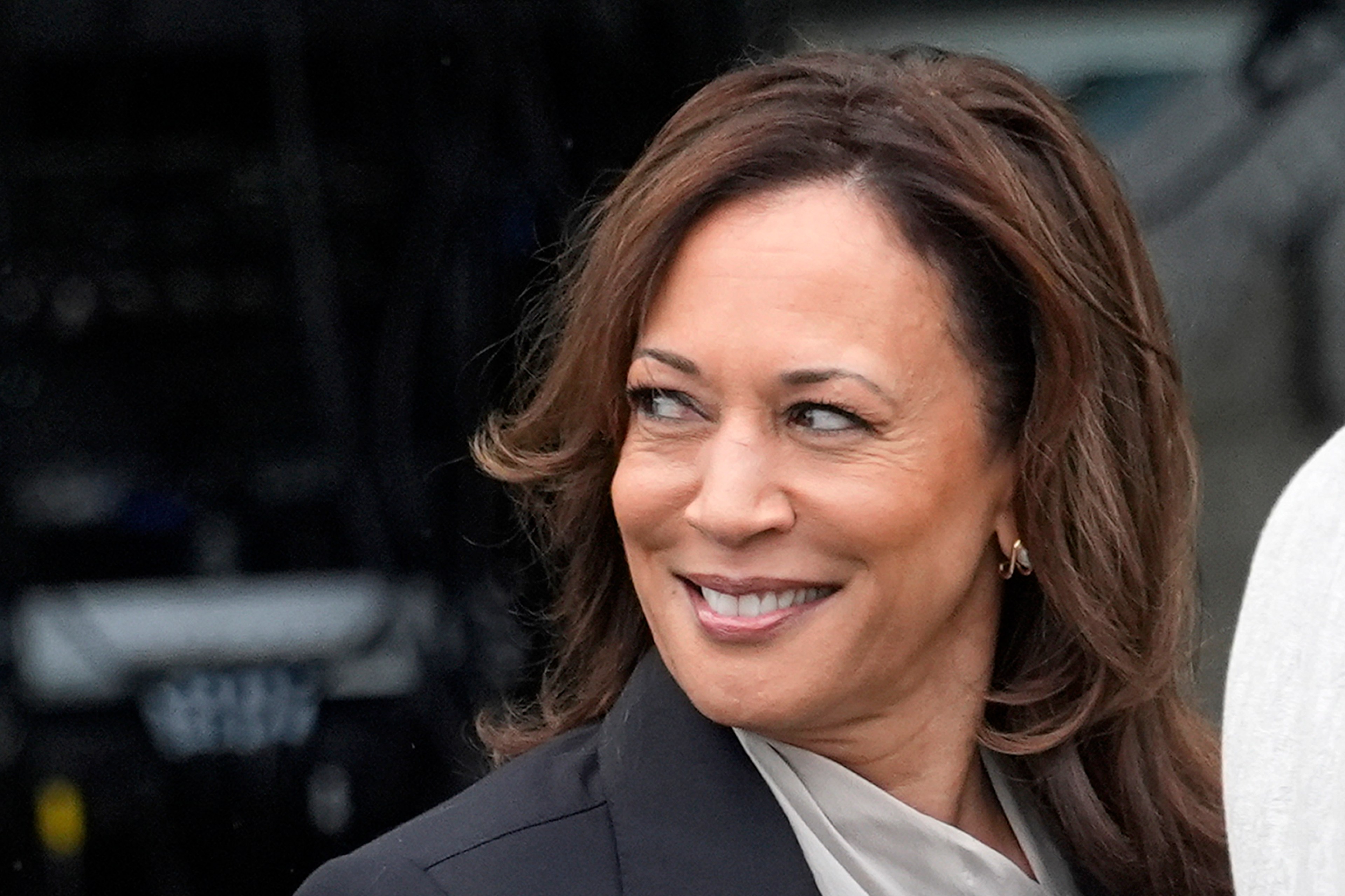 A woman with shoulder-length brown hair smiles while looking to her left. She is wearing a black blazer and has small hoop earrings. The background is indistinct.