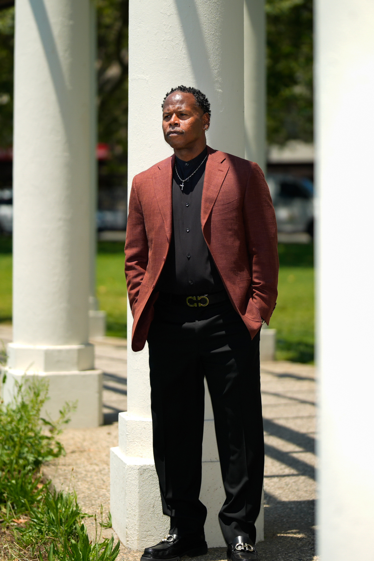 A man stands by white columns, wearing a rust-colored blazer, black shirt, and black pants with a stylish belt. He has his hands in his pockets and looks pensive.